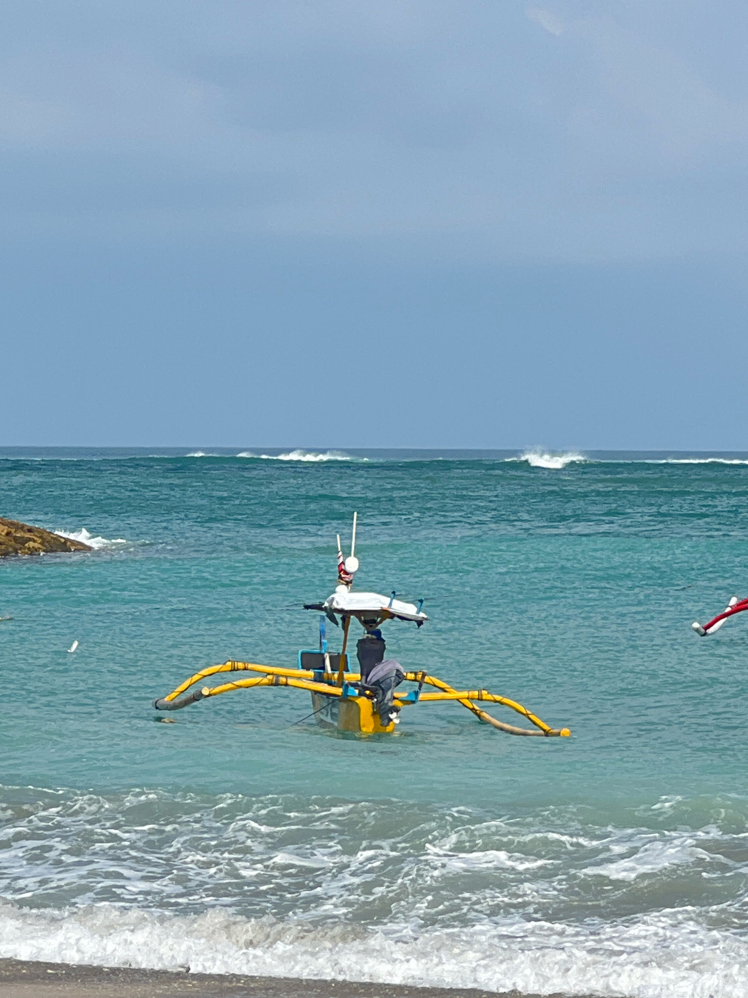 A yellow and blue traditional Balinese fishing boat, also known as a jukung, is anchored in the turquoise waters off the coast of Kuta, Indonesia. The boat features outriggers painted bright yellow and has a small motor attached to the stern. A person, wearing dark clothing, is seated in the boat, tending to equipment. In the distance, waves are breaking on the horizon, indicating a moderate surf. The sky above is a clear, pale blue. On the left side of the frame, a rocky outcrop juts out into the water. The foreground shows the gentle wash of waves lapping against a sandy shore. Another jukung with a red and white flag is partially visible on the far right of the image. The scene captures a typical coastal view in Kuta, known for its beaches and surf culture.