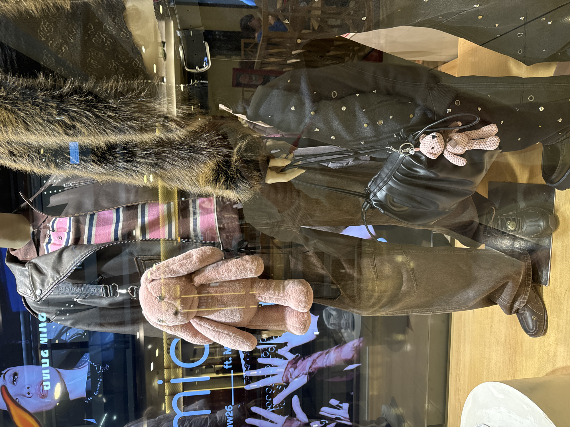 A photograph taken in Buenos Aires, Argentina, depicts a mannequin displayed in a store window. The mannequin is dressed in a brown and pink striped shirt, a black leather jacket with a brown fur collar, and brown trousers, with a black handbag and black shoes completing the ensemble. A pink stuffed bunny is attached to the jacket. The window has reflections of the environment and posters.