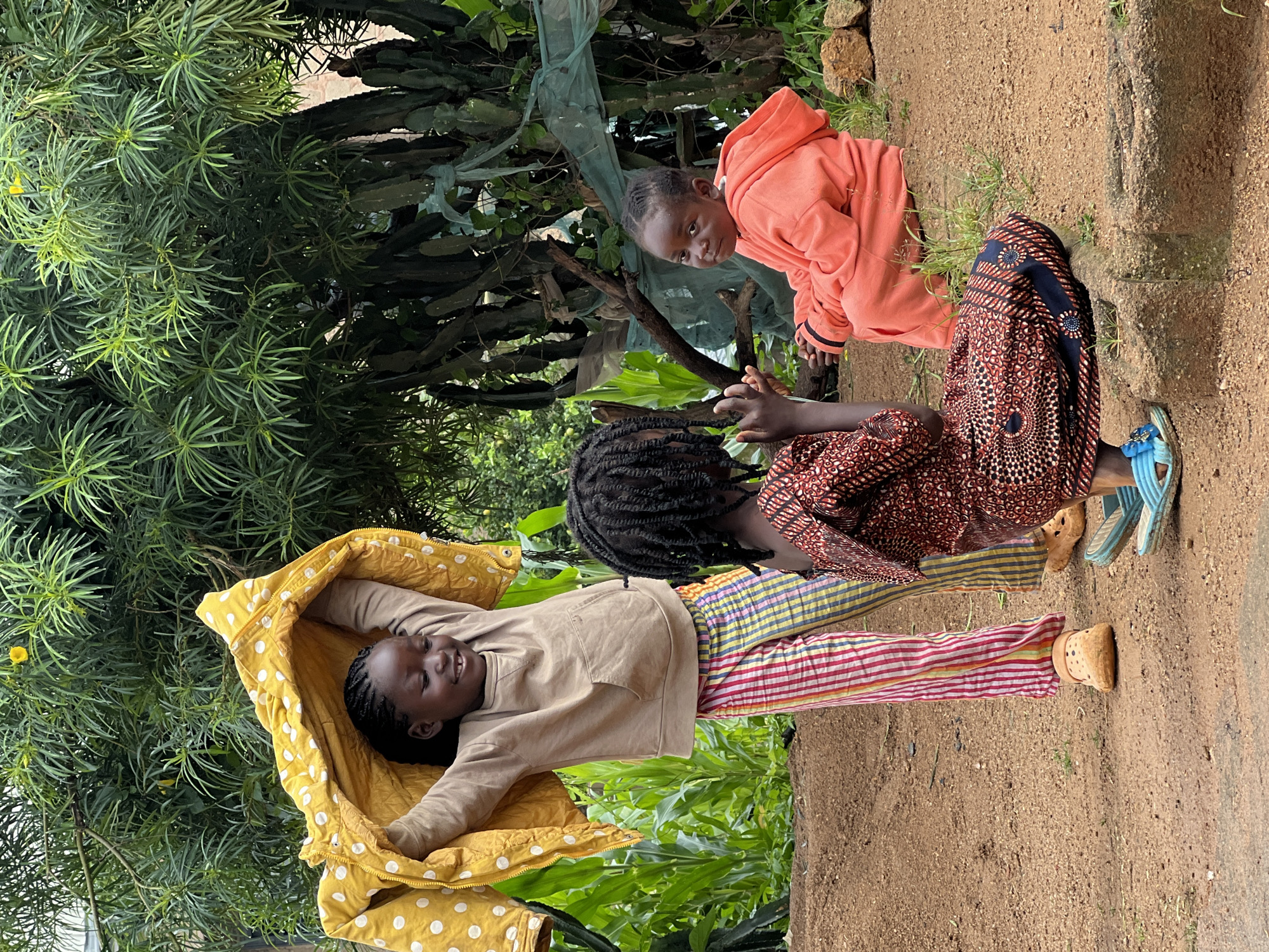 Three young Black girls are interacting outdoors in an area with dirt ground and lush green foliage. The girl on the left, wearing a tan hooded sweatshirt and colorful striped pants, holds a yellow blanket with white polka dots above her head. The girl in the middle, facing away from the camera, wears a patterned red and black dress. The girl on the right, wearing an orange hooded sweatshirt, is seated on the ground facing the other two. In the background, there are large green plants, including what appear to be cactus-like plants, and some visible fencing. The scene suggests a casual outdoor gathering. The presence of lush vegetation and the overall environment are consistent with the climate and landscape found in Jos, Nigeria.