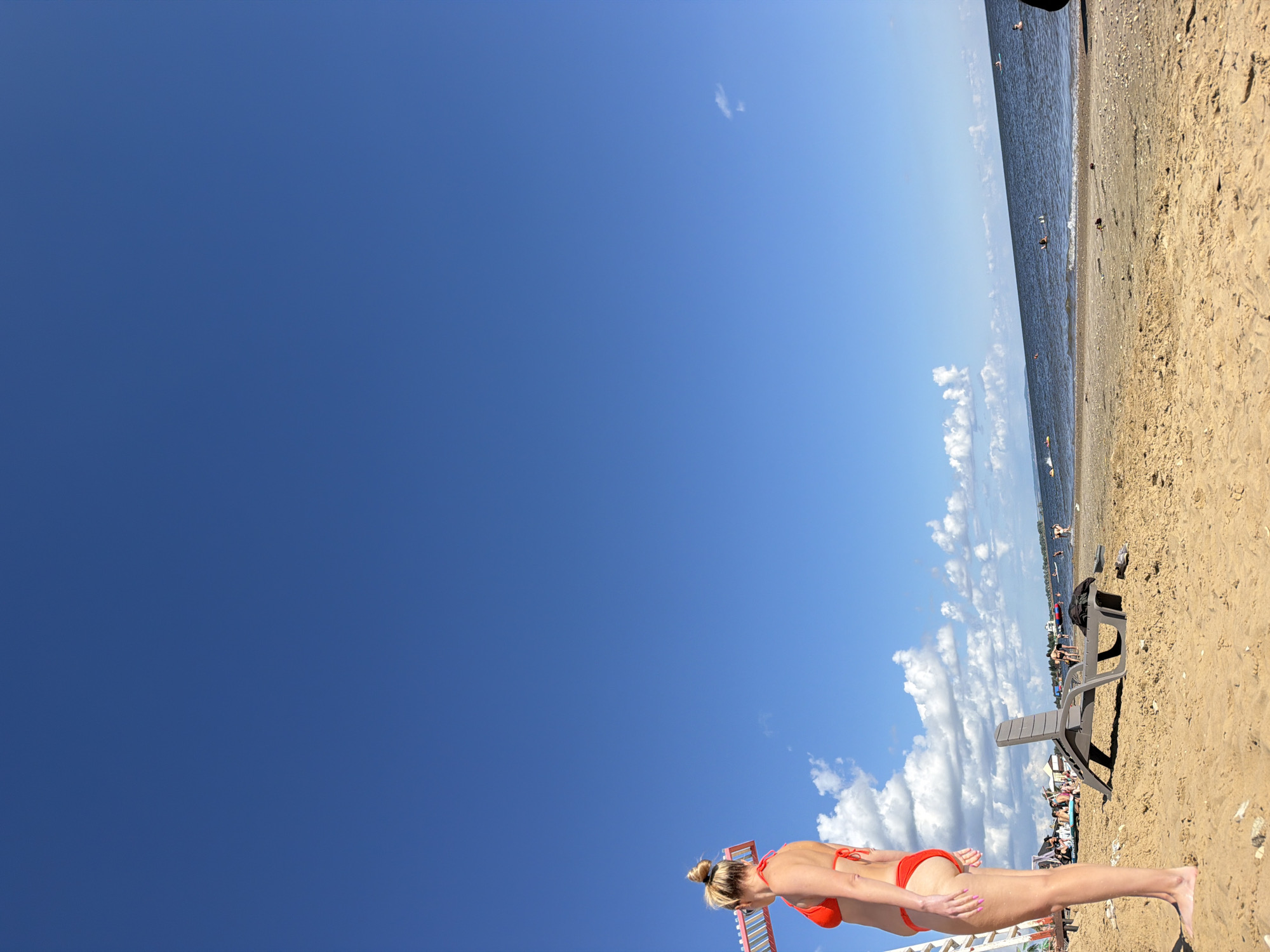 An outdoor, eye-level, full shot captures a sunny day at a beach. The sky is a vibrant blue with scattered white clouds. In the foreground, a woman with blonde hair pulled into a bun, wearing a red bikini, stands facing left. She is partially in the frame, with her back to the camera. To her right, a grey plastic lounge chair is positioned on the sandy beach. Further back, the water meets the shore, with several people swimming and playing in the ocean. In the distance, there are buildings and what appears to be a pier or a long structure extending into the water. The overall scene depicts a lively beach day with people enjoying the water and sun.