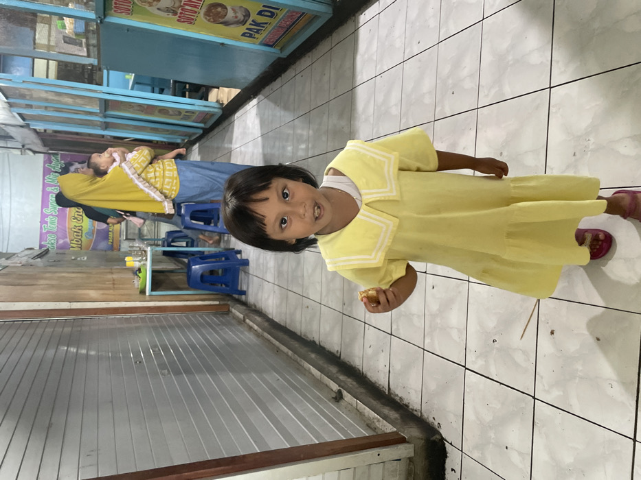 An indoor or semi-enclosed market alleyway in Nganguk, Indonesia, features a white tiled floor with dark grout lines. In the foreground, a young child with dark, straight hair, wearing a yellow short-sleeved dress with a white sailor collar and pink sandals, stands facing the camera. The child holds a small, light-brown, irregular food item in her right hand and has an open-mouthed expression. A thin, dark stick object lies on the floor near her feet. In the midground, to the left, a light-blue square table with two blue plastic stools is positioned against a wall with a closed corrugated metal security shutter and wooden paneling above it. To the right, a series of light-blue framed food stalls are visible, displaying Indonesian text and images, including signs for "Bakso Tenis Super & Mie Ayam" and "SOTO AYAM PAK DI." Further in the background, a woman wearing a yellow hijab and a patterned top over a blue skirt holds an infant, who is dressed in light clothing and faces away from the camera. Another woman in a dark shirt is partially visible standing near the table. The area is illuminated by overhead artificial lighting.