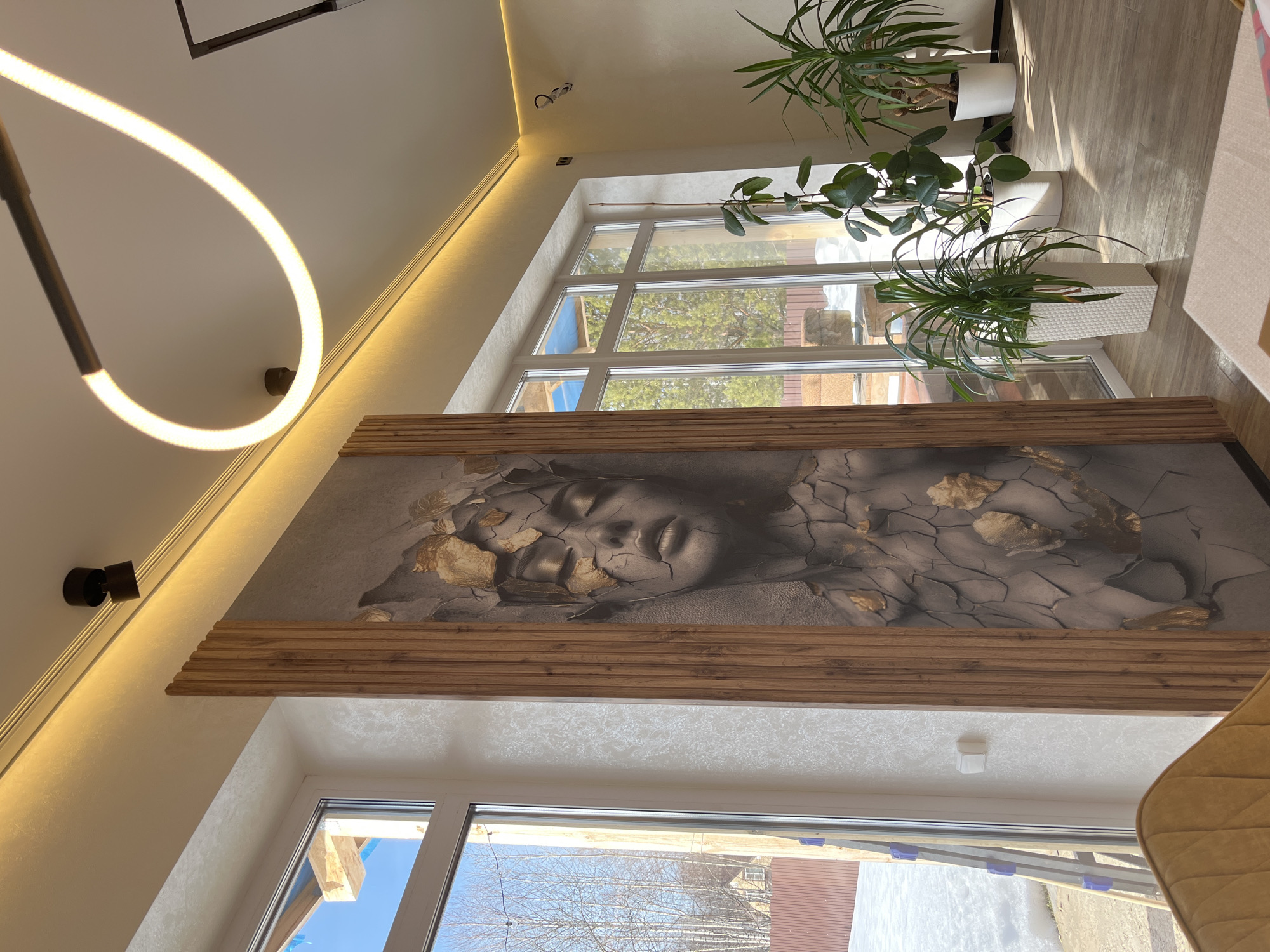 The image displays an interior room located in Новая Чернушка, Russia, overlooking an exterior winter scene. A large, vertical wall mural occupies the central portion of the main wall, depicting a woman's face with closed eyes, rendered in a grey tone with cracked textures and scattered gold leaf-like elements. The mural is flanked on its right by a section of vertical wooden slats. To the left of the mural, a large, multi-pane window with white frames offers a view of snow-covered ground, numerous bare trees, a reddish-brown fence, and a distant residential structure. A silver ladder with blue accents is visible leaning against the exterior wall outside this window. A white electrical switch is mounted on the wall adjacent to the left window. The room features a light-colored ceiling with perimeter LED strip lighting, a black recessed spotlight, and a distinct curved white LED light fixture. The flooring consists of light-toned wood planks. Several potted green plants are arranged near the windows on the right, housed in white rectangular and cylindrical planters. A segment of a mustard-yellow upholstered chair is visible in the lower-left foreground, and a light-colored rug partially covers the floor in the lower-right. No individuals are present in the scene.
