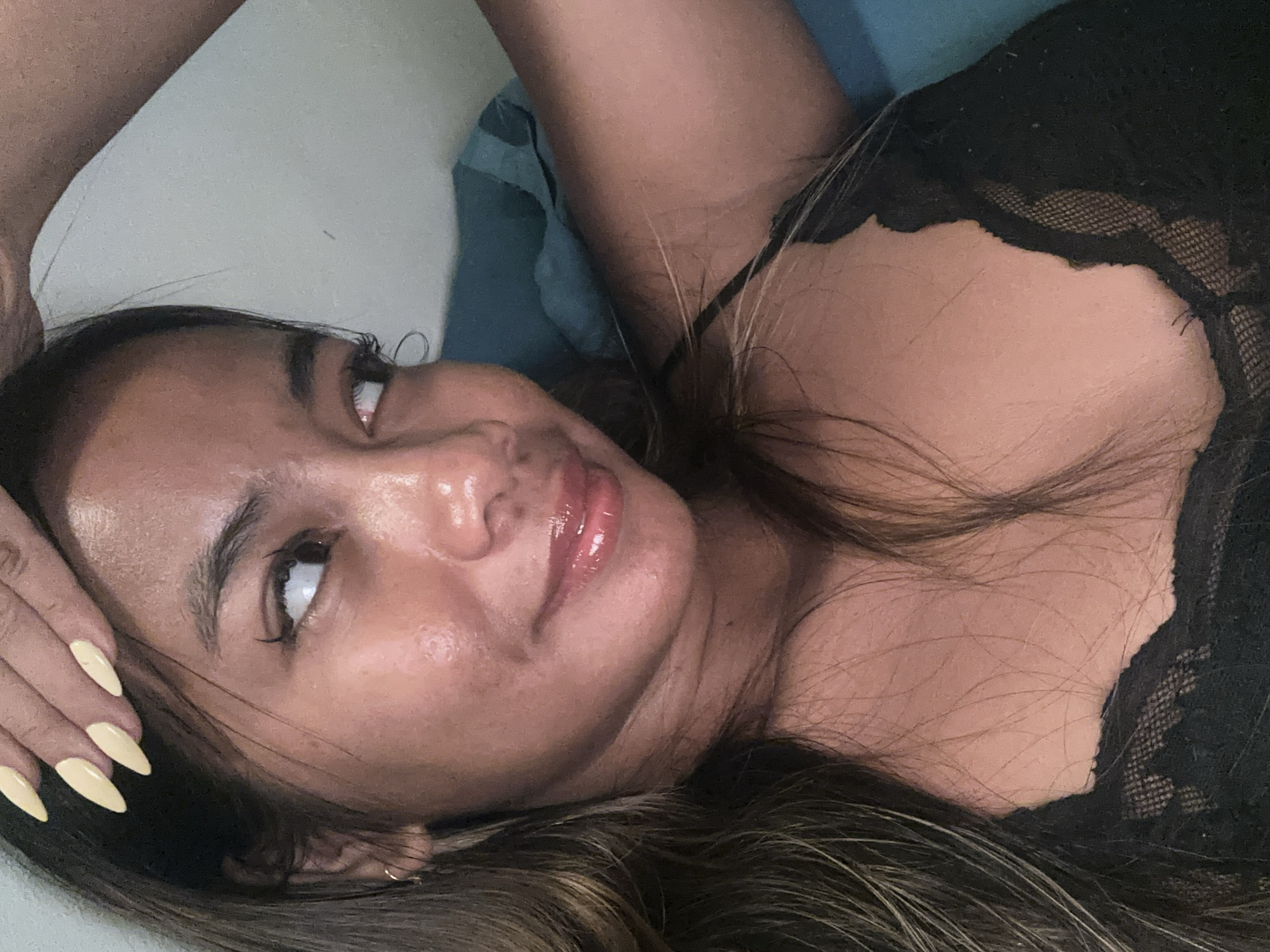 This media file features a close-up, low-angle shot of a young woman lying down. She has long, dark brown hair that cascades around her face and shoulders. Her eyes are looking upwards, and her lips are slightly parted, with a sheen of lip gloss. She is wearing a black lace top. Her left hand is resting on her head, with her fingernails painted a pale yellow. The background is out of focus, appearing to be a blue surface, possibly a pillow or bed. The lighting suggests it is daytime, indoors, with a warm glow on her skin, highlighting a dewy complexion. The overall mood is intimate and relaxed. No text is visible in the image.