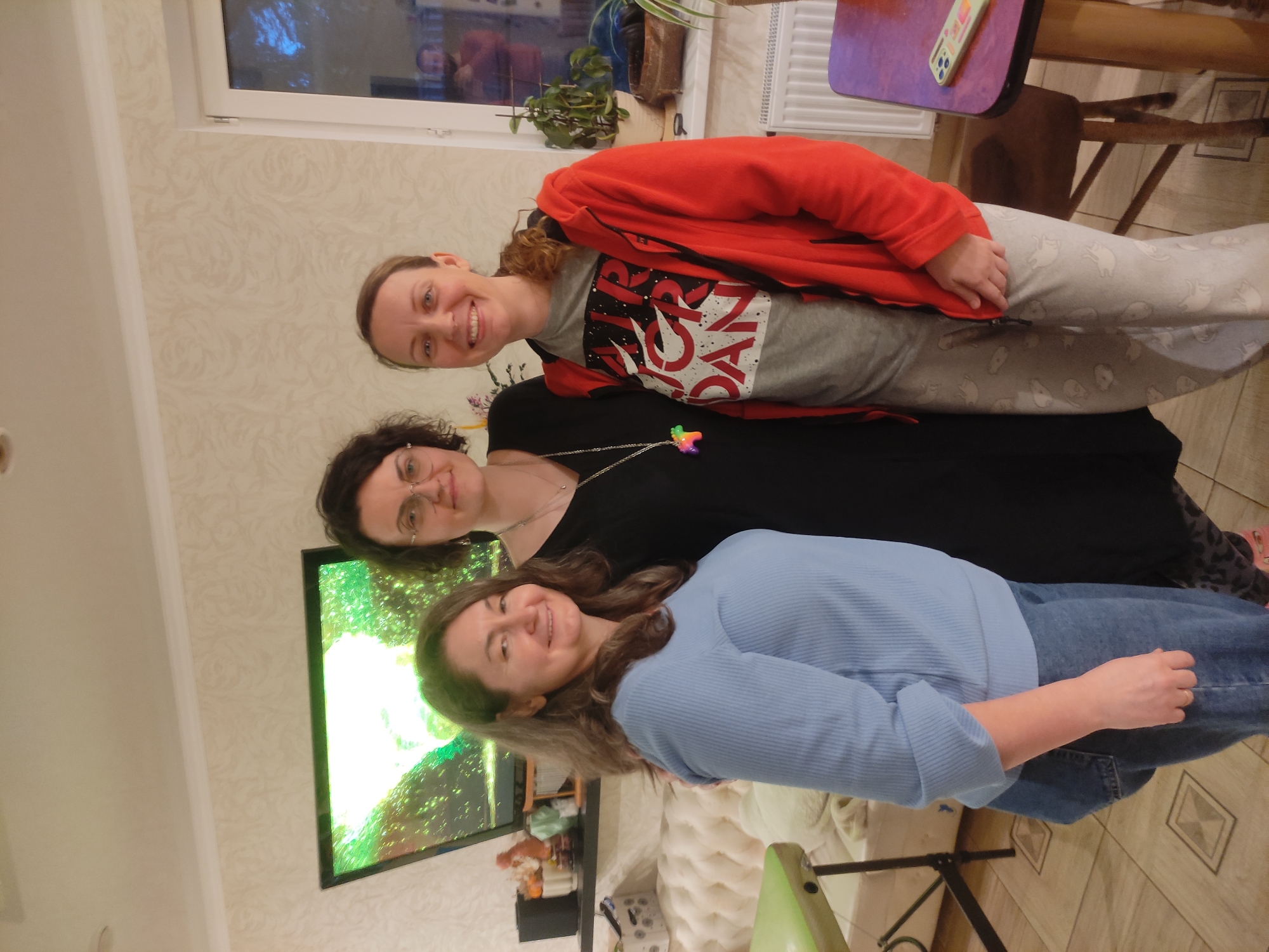 Three women are posing for a photo in an indoor setting.
The woman on the top is wearing a red jacket over a grey t-shirt and patterned grey pants.
The woman in the middle is wearing glasses and a black dress with a rainbow-colored pendant.
The woman in the bottom is wearing a light blue shirt and jeans.
Behind them, there is a TV screen displaying a green image, a window with plants on the sill, and furniture including a table and a chair. The background features patterned wallpaper. The location is in the city of Unknown, Unknown.