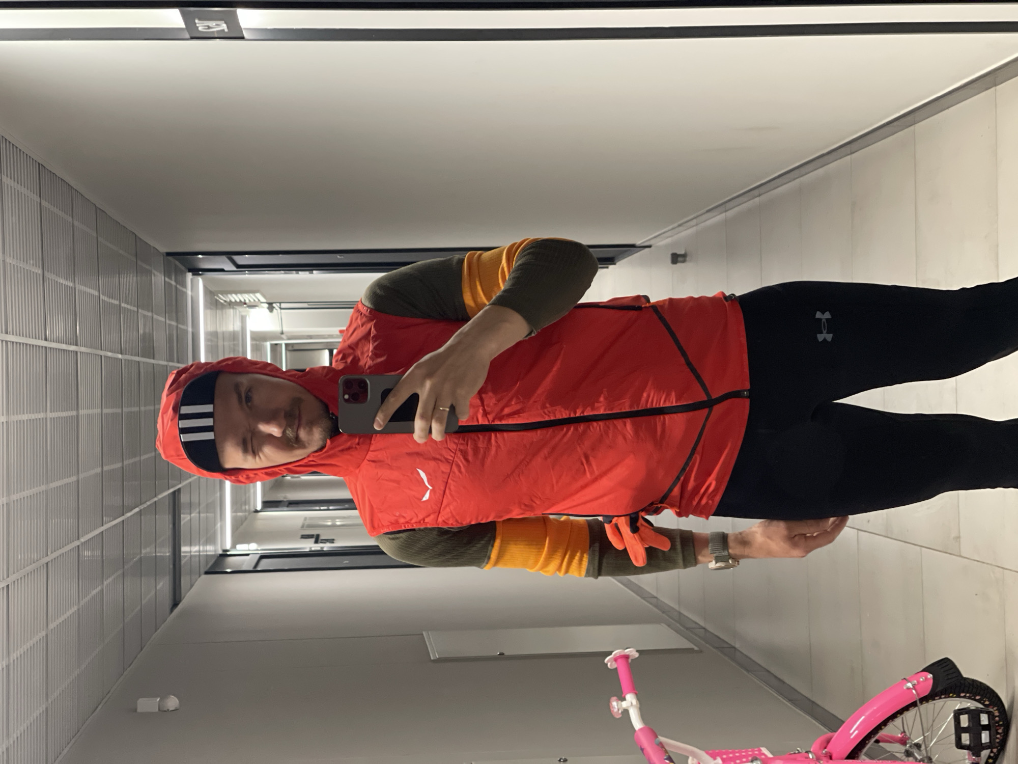 A man is standing in a hallway, taking a selfie in a mirror. The hallway has white walls with horizontal grooved paneling. The floor is off-white and clean. Overhead lighting runs along the length of the hallway. The man is wearing a red hooded vest over a long-sleeved shirt with olive green and orange stripes on the sleeves. He has black leggings with an "Under Armour" logo on the left thigh. He also has a black headband with white stripes and is holding a smartphone. A pink child's bicycle is in the foreground on the right. The location is not identifiable as Izhevsk, Russia.