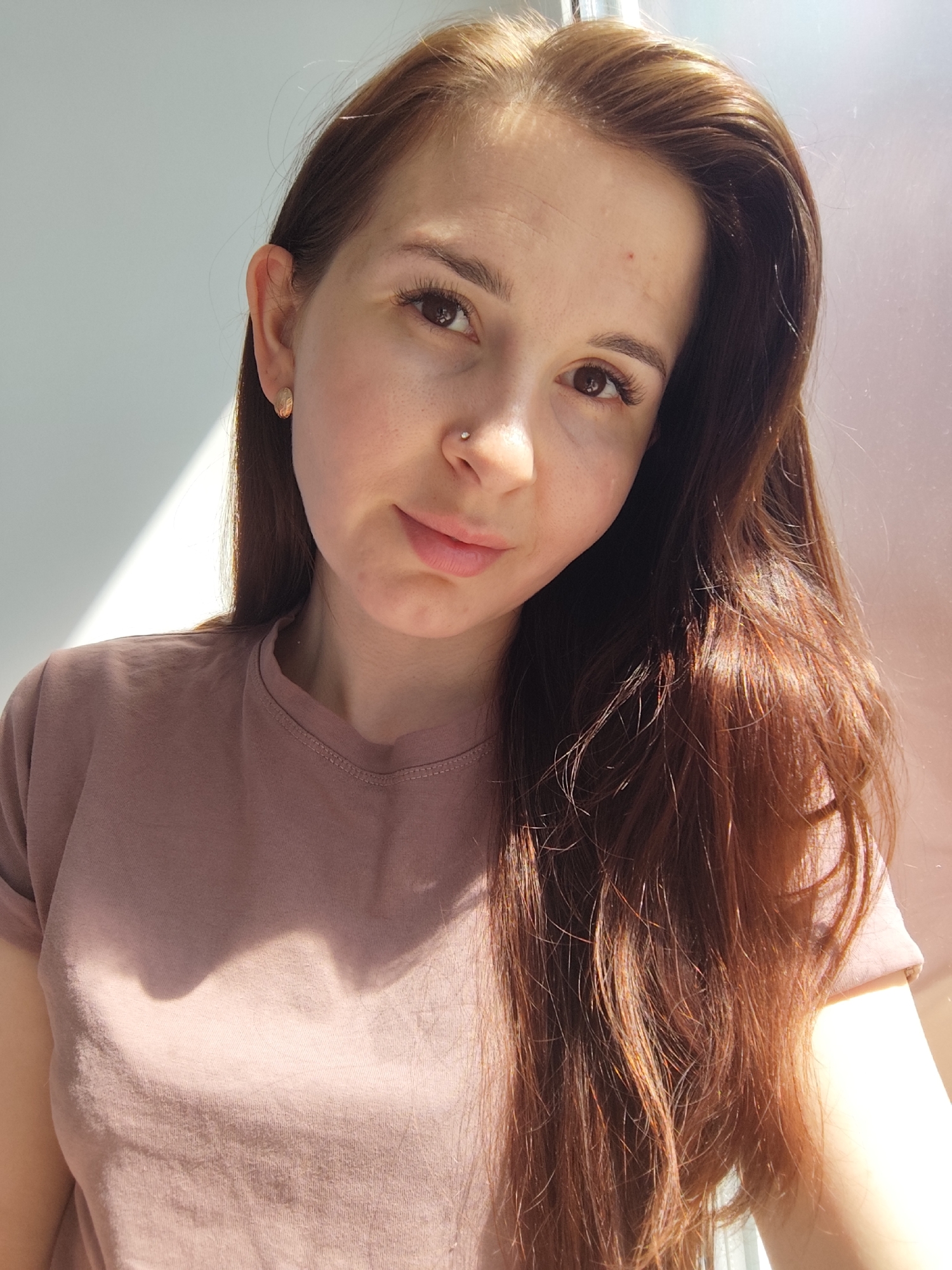 A woman with light brown hair and fair skin is posing for a photo. She is wearing a light brown t-shirt and has a small stud in her nose. Her hair is parted to the side and falls over her shoulders. The light source is strong, creating shadows on her face and t-shirt. The background appears to be a white wall. The location is unknown, but the city of Фоки, Russia was mentioned. There is no information to confirm the location within the photograph.