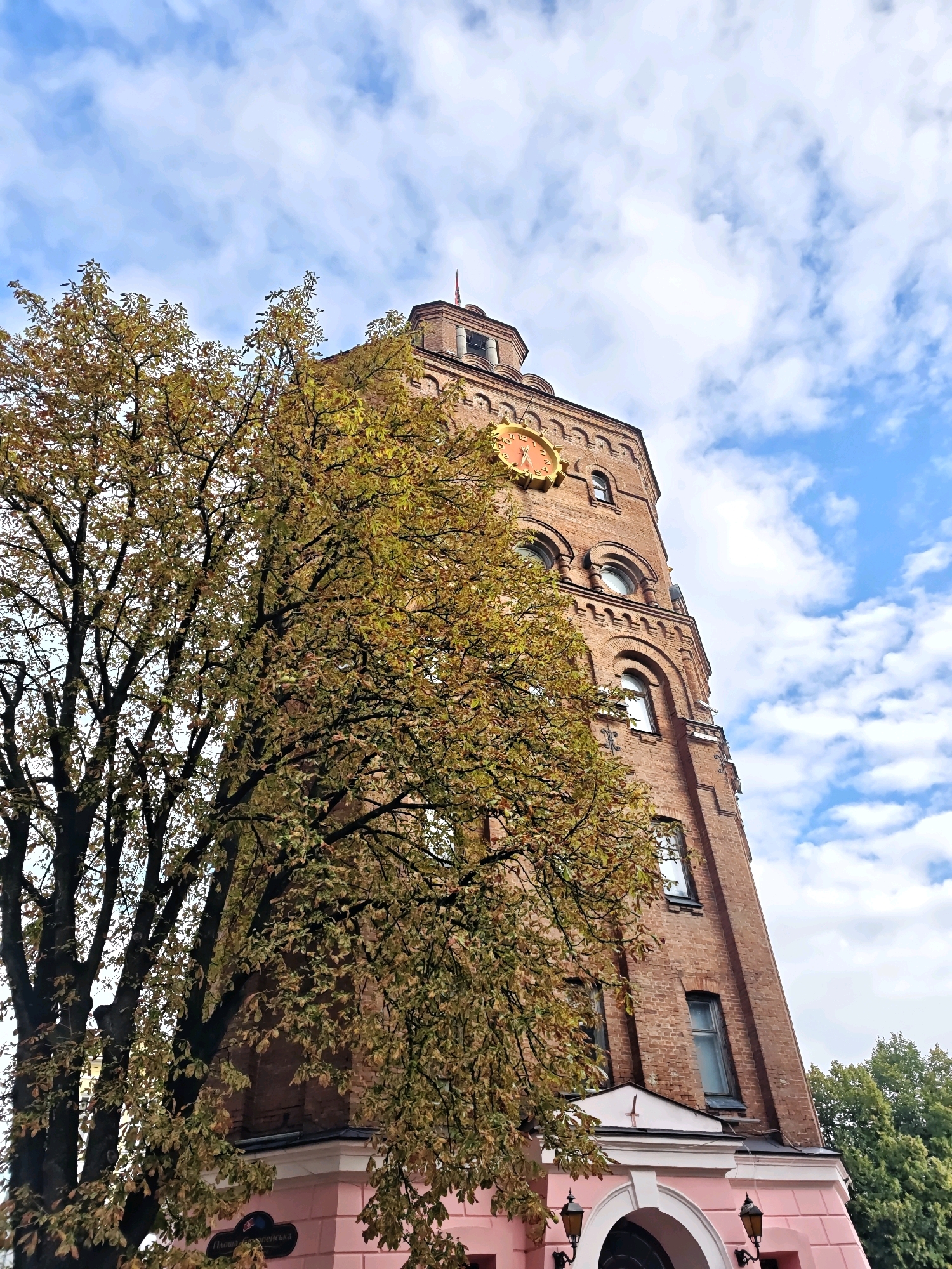 The image shows a tall, red brick building with a prominent clock face, likely a historic tower or part of a larger structure. The building has arched windows and ornate architectural details. A large tree with yellowing leaves, suggesting autumn, is partially obscuring the building on the left side. The sky is blue with scattered white clouds.
This building is identified as the **Water Tower on Postova Street in Vinnytsia, Ukraine**. It is a significant landmark in the city, often referred to as the "Kvitka" (Flower) due to its unique architectural style. The tower was built in 1910 and served as a water supply facility. It has since been converted into a museum and a viewing platform. The surrounding area appears to be urban with some greenery visible in the background. There are no people or animals visible in the image.