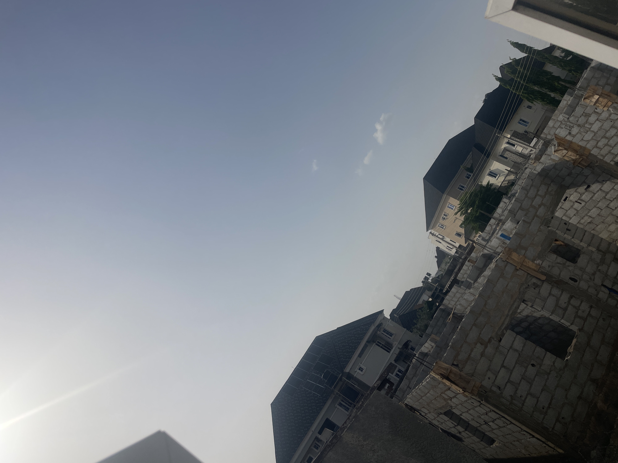 The image captures an outdoor scene showcasing urban development, likely in Dushepe, Nigeria, with a clear, hazy sky dominating the upper frame. The photograph is taken at an oblique angle, tilting the horizon line.
The sky is a pale blue, suggesting a bright day, though a noticeable haze is present. A few sparse, wispy white clouds are scattered across the upper right. Strong sunlight causes a bright glare originating from the lower-left corner of the image, indicating either morning or late afternoon.
In the foreground, on the right side, an unfinished multi-story building is under construction, characterized by its grey concrete block walls. Several rectangular openings are visible, indicating future windows or doors, and some wooden supports or formwork are integrated into the structure.
Beyond the construction site, in the midground, stand several completed residential buildings. These are substantial multi-story houses featuring dark, possibly tiled or shingled, sloped roofs and light-colored (likely white or beige) exteriors. Tall, slender green trees are visible behind some of these finished properties, adding natural elements to the built environment. Thin utility lines stretch across the midground, indicating established infrastructure.
No people are visible in the scene, and there is no discernible text. The image portrays a state of ongoing development and existing residential structures under clear, sunny, yet somewhat hazy, daytime conditions.