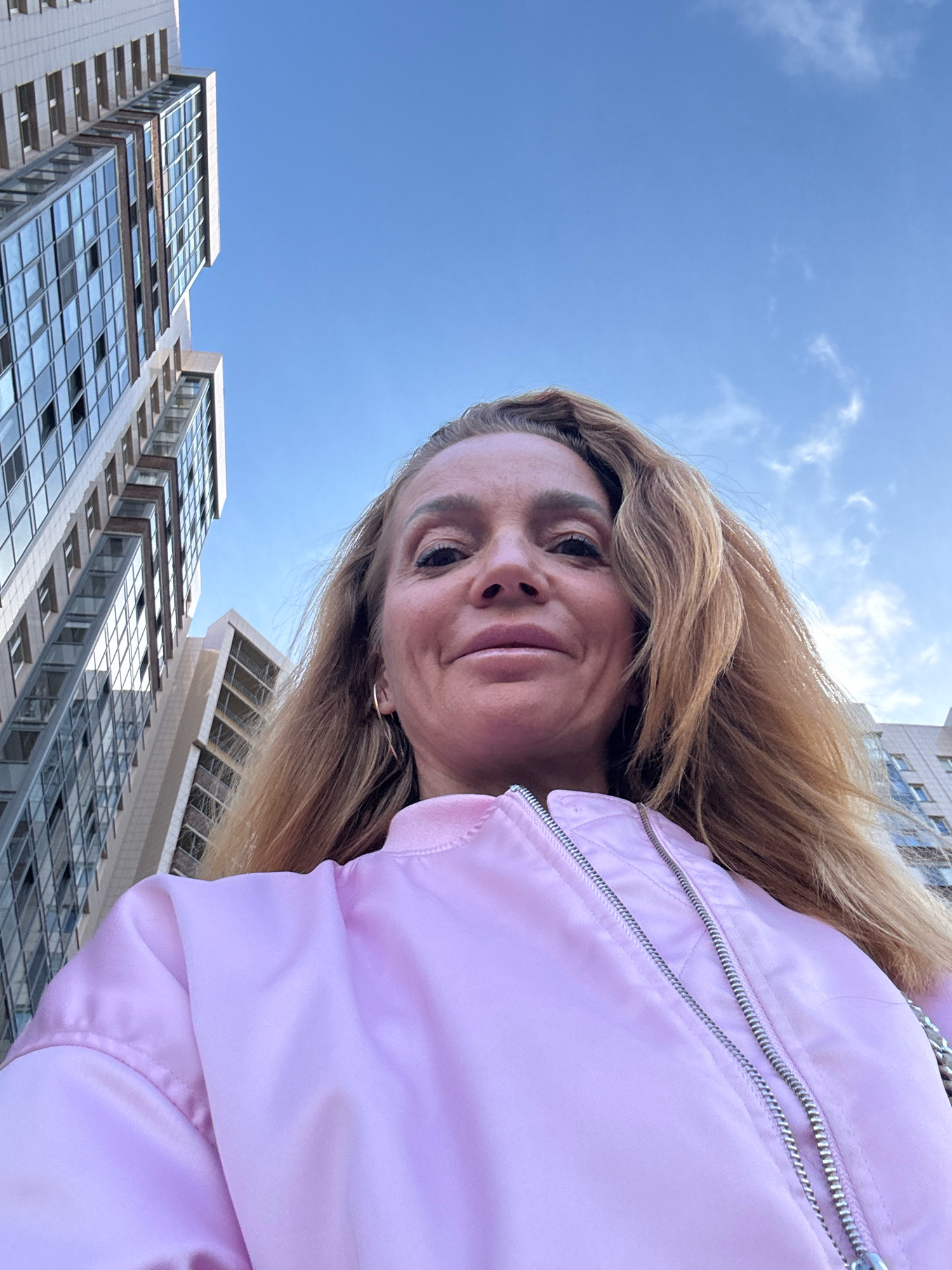 A low-angle, selfie-style shot shows a woman in a pink bomber jacket, with buildings and a blue sky in the background. The woman has blonde hair and is looking at the camera. The buildings appear to be modern high-rise apartment buildings. The image was taken outside in an urban environment.