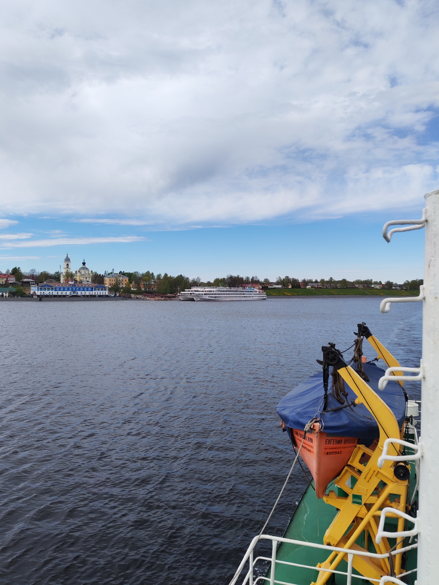 The image depicts a view from a ship on the Volga River, likely in the vicinity of the city of Myshkin, Russia. In the distance, a cityscape is visible, featuring distinctive architecture with a white church and a building with a blue facade, characteristic of Myshkin. Another white ship is docked alongside the city. In the foreground, part of the ship from which the photo is taken is visible, including a lifeboat covered with a blue tarp and secured by yellow davits and ropes. The water of the Volga River fills the middle ground. The sky above is blue with scattered white clouds.