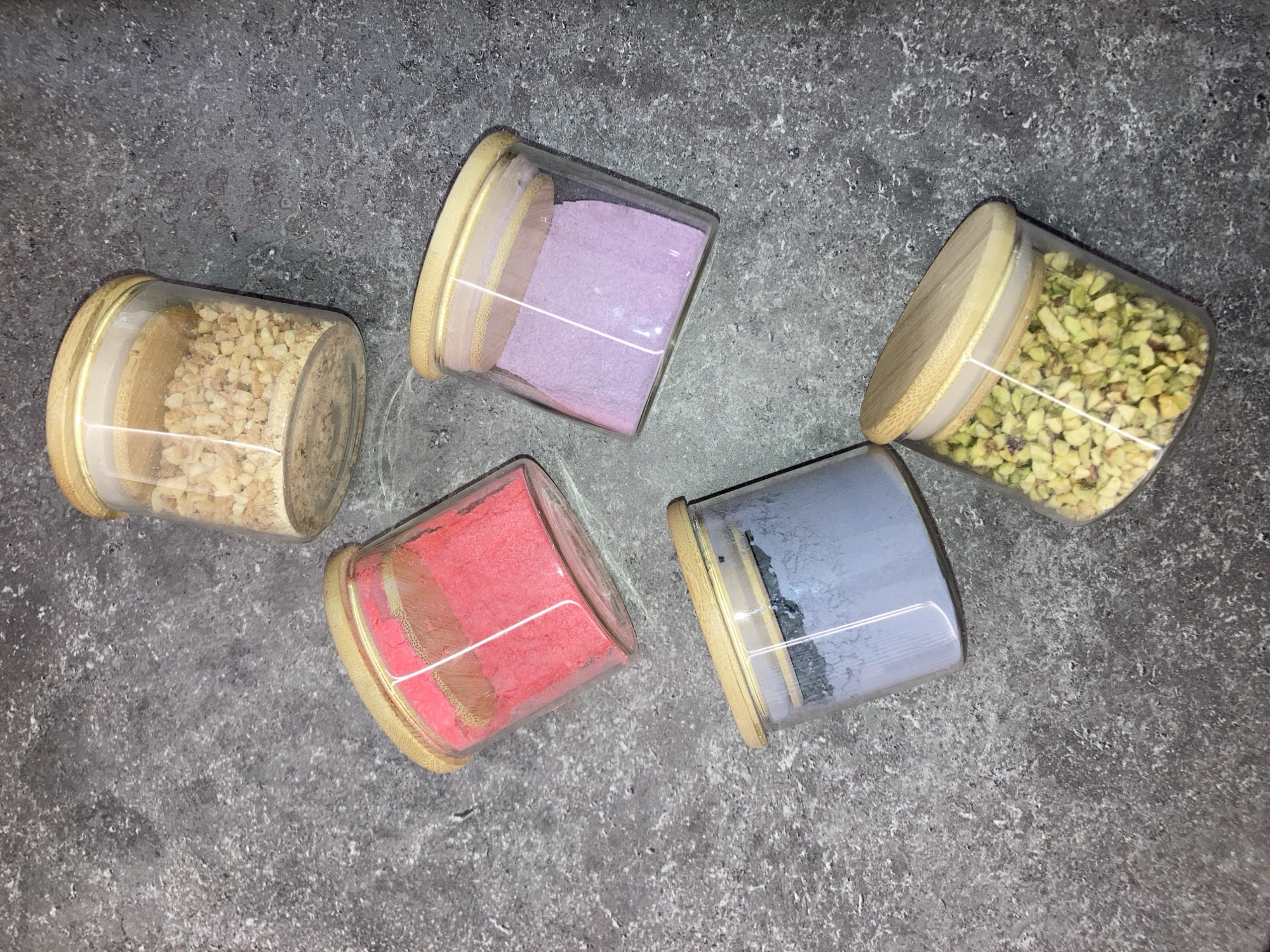 The image shows five cylindrical glass jars with wooden lids arranged on a gray, textured surface. Each jar contains a different colored substance: one contains chopped nuts, another a purple powder, a third chopped pistachios, a fourth a pink powder, and the last a grayish-blue powder. The lighting appears diffused, casting subtle shadows. The scene is not explicitly identifiable to any location in Chaikovsky, Russia, without additional context or information.