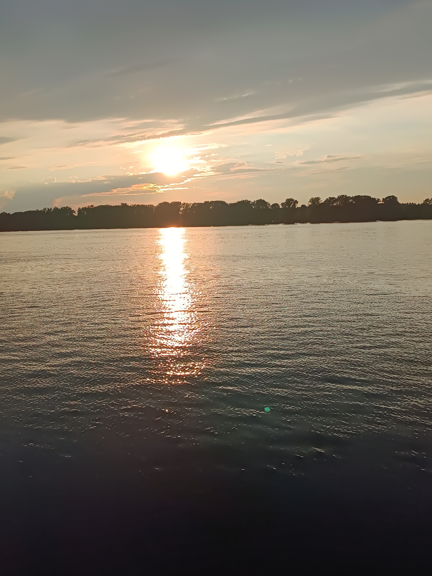 A vertical, eye-level, wide shot captures a sunset over a wide body of water, likely the Kama River near Chaikovsky, Russia. The sun, a bright, concentrated orb, is visible in the upper center of the frame, partially obscured by light clouds. Its rays create a shimmering, golden reflection that stretches across the water's surface towards the viewer. The water itself is rippled, with darker areas in the foreground and mid-ground, suggesting depth and possibly movement. The far bank of the river is lined with a dense silhouette of trees, their dark forms stark against the lighter sky. The sky above is a mix of soft grays and pale oranges, with wispy clouds catching the dying light. No people or specific man-made objects are clearly discernible in the scene. The overall impression is one of natural beauty and tranquility at the close of day.