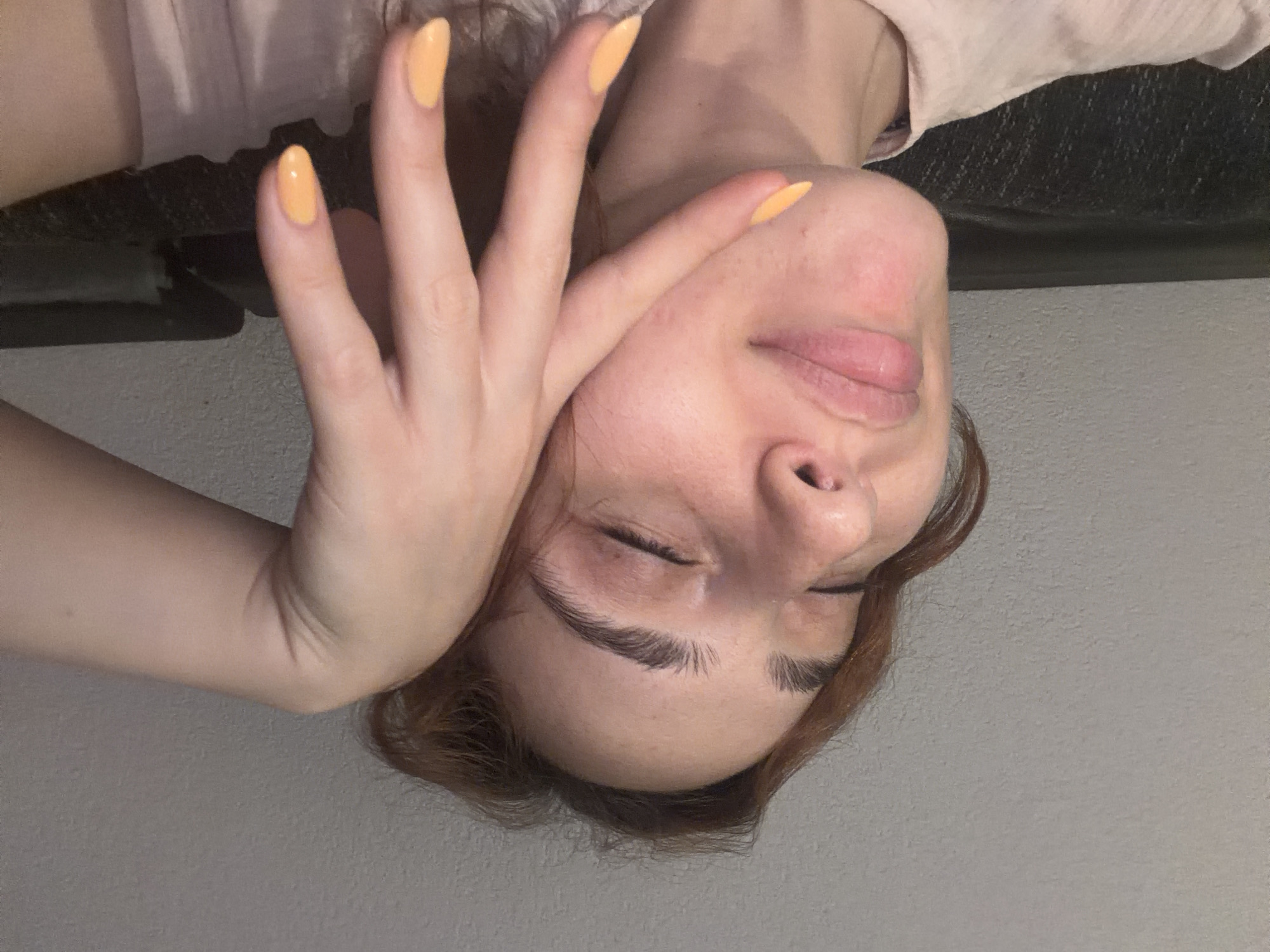 This is a close-up, upside-down, vertical photograph of a person's face and hand. The person has light skin, brown hair, and is wearing a light pink shirt. Their eyes are closed, and their lips are slightly parted. The hand, with bright yellow nail polish on the fingernails, is placed across the person's face, covering their nose and mouth area. The background appears to be a textured wall. The lighting suggests an indoor setting, possibly during the day. No text is visible in the image. The overall mood conveyed is relaxed or introspective.