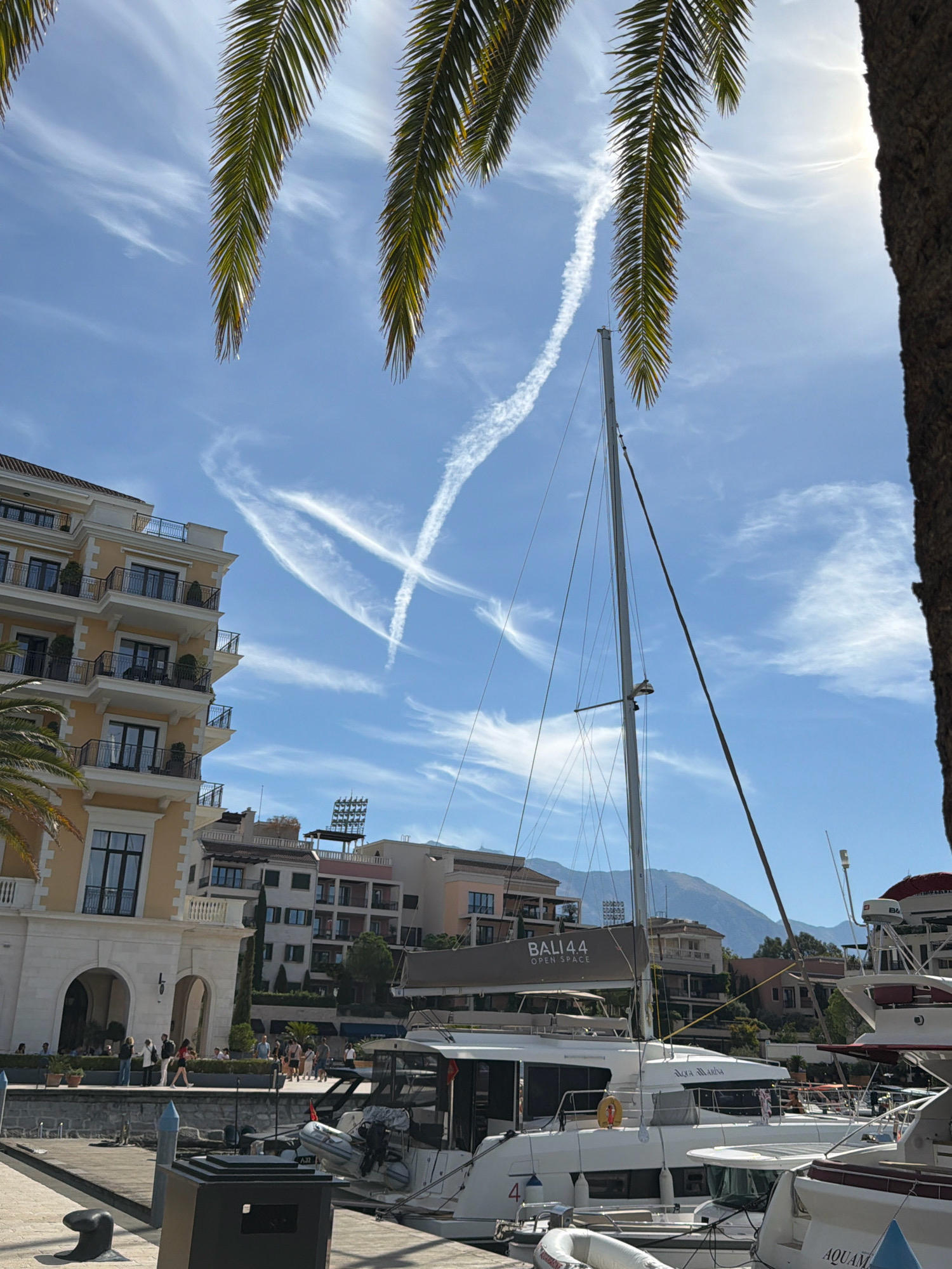 In Tivat, Montenegro, a clear sky with contrails forms a prominent "X" over a marina. Palm fronds partially obscure the view of a modern, multi-story yellow building with balconies on the left. In the foreground, a white catamaran named "BALI 44 OPEN SPACE" is docked, with another white boat, possibly named "Aqua Marina," beside it. The marina features a paved walkway with several people strolling. In the background, more buildings and mountains rise under the bright sun.