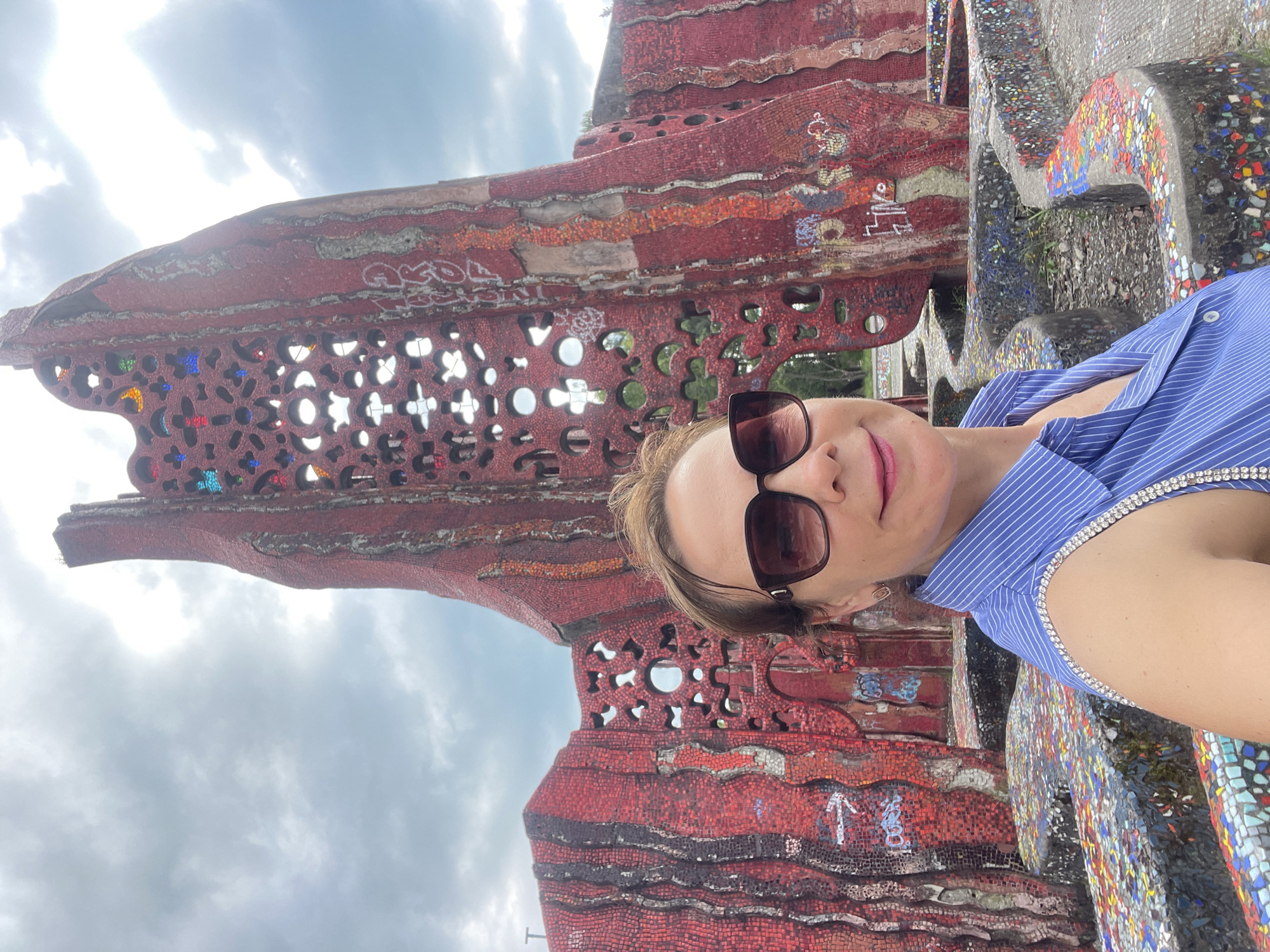 A woman takes a selfie in front of a large, red, mosaic-covered structure. The structure is abstract and appears to be part of an outdoor art installation or park. It features intricate patterns and cutouts, some of which are filled with colored glass. The woman is wearing a blue and white striped shirt with rhinestone embellishments on the shoulders and large sunglasses. She has brown hair and is smiling. The background shows a cloudy sky and some greenery. The location is identified as Sochi, Russia, likely within the "Dreamland" park.