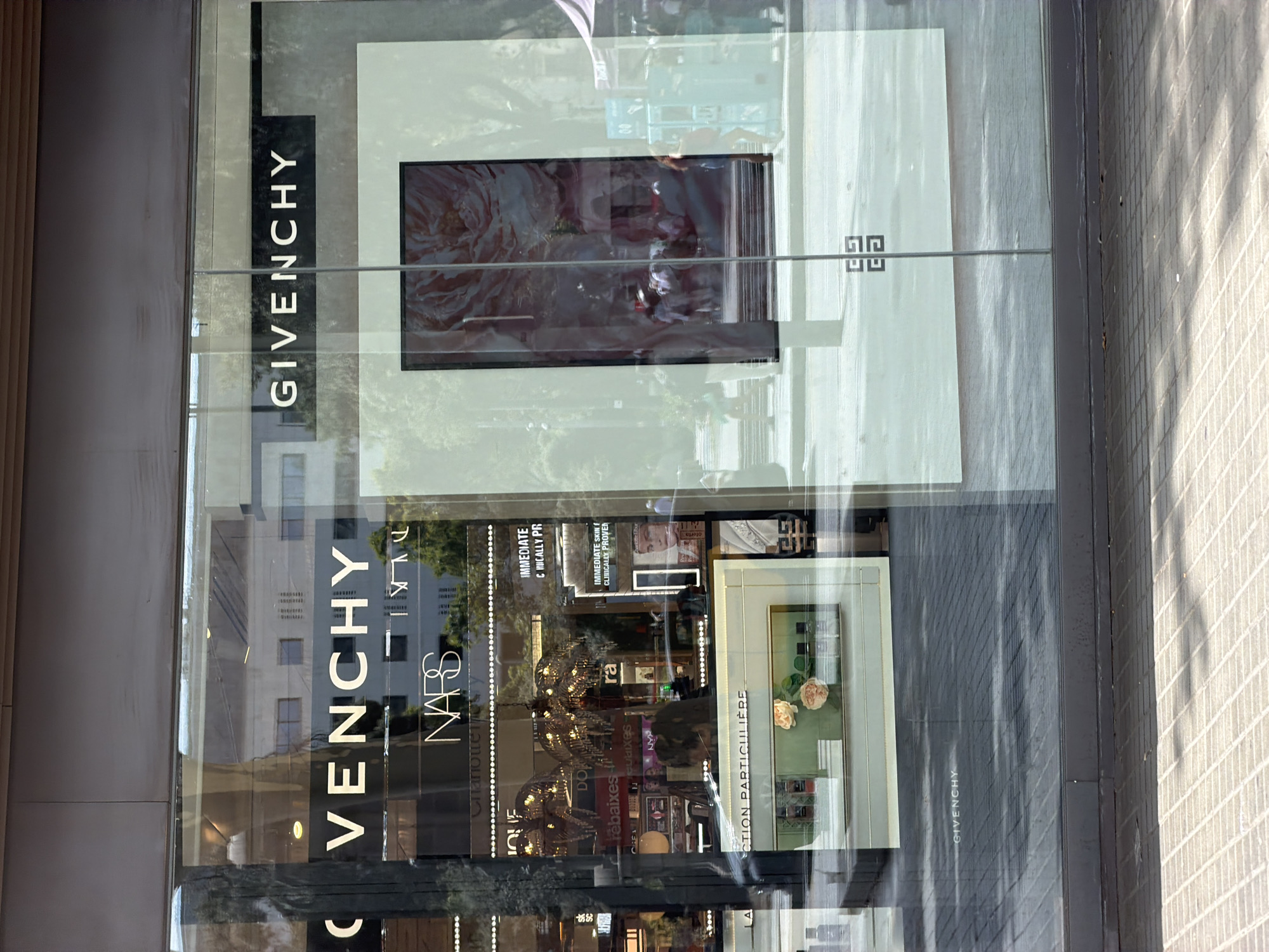 The image captures the exterior of a Givenchy store, likely located on a commercial street in Barcelona, Spain. The store features a large glass facade that reflects the surrounding environment. Prominently displayed on the facade are the brand names "GIVENCHY" and "NARS." Inside the store, the reflection reveals a display area with various cosmetic products, including perfume bottles and makeup. Two palm tree-shaped chandeliers with crystal embellishments are visible hanging from the ceiling. A digital screen displaying a floral image is positioned within the store. The street outside appears to be paved with patterned tiles, and the reflection shows people walking on the sidewalk. The overall scene depicts a high-end retail establishment in a bustling urban setting.