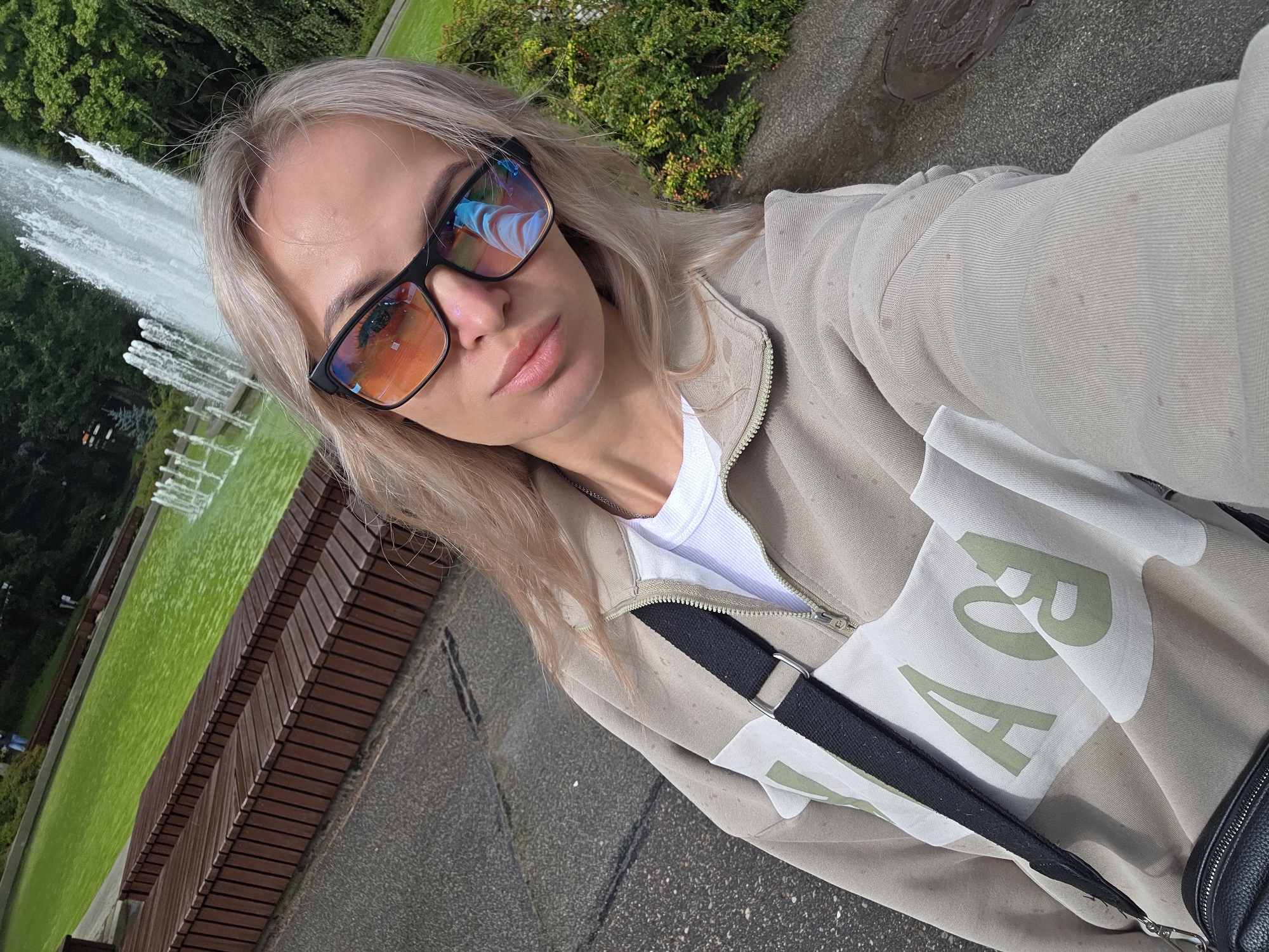 In a park setting in Kaliningrad, Russia, a young woman with blonde hair and sunglasses is taking a selfie. She is wearing a light-colored quarter-zip sweatshirt over a white t-shirt and has a black strap from a bag across her chest. In the background, a large fountain sprays water into a bright green pond, surrounded by trees and wooden benches. The ground is paved with grey cobblestones.