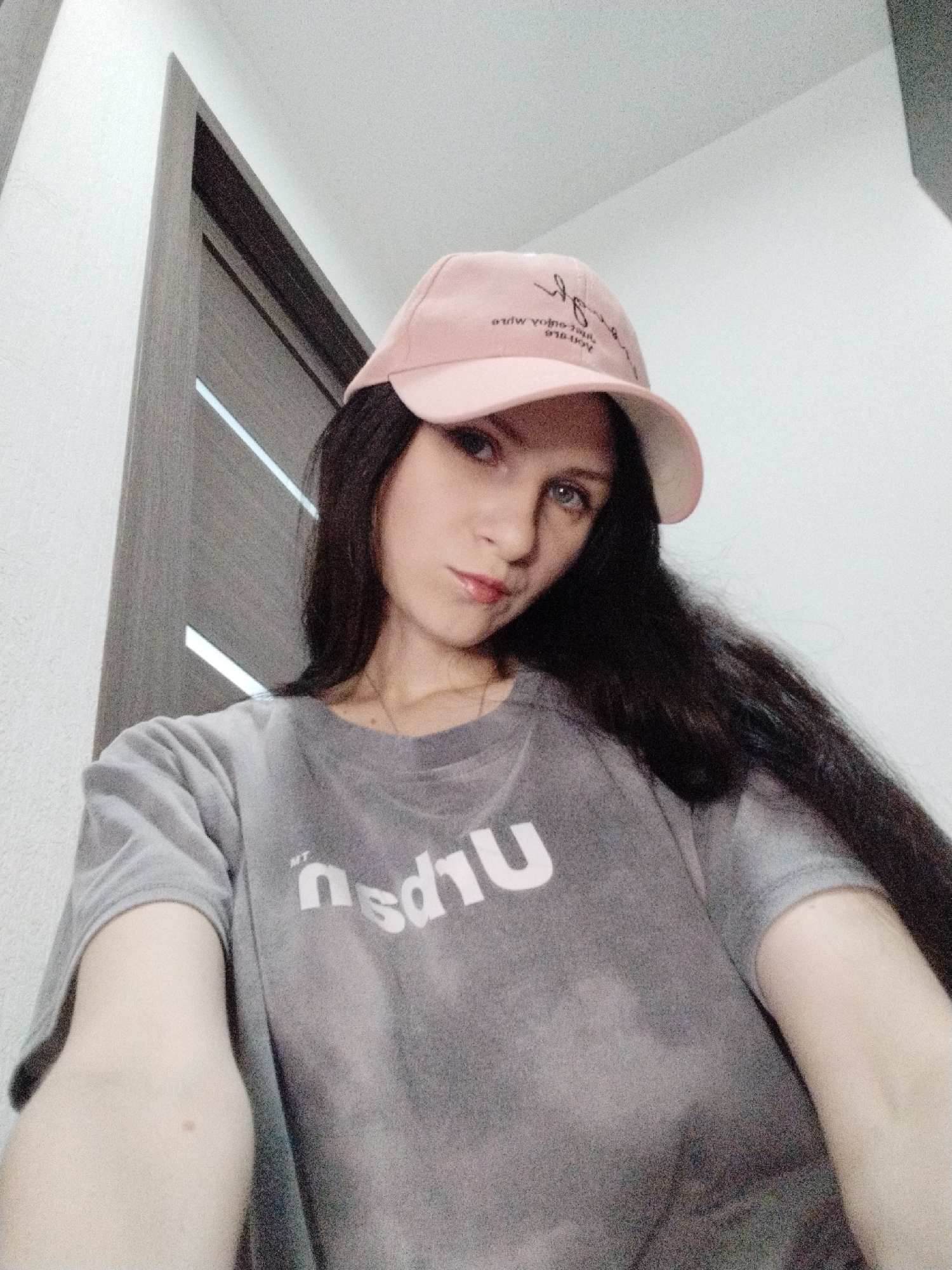A young woman with long, dark hair and light blue eyes is looking at the camera. She is wearing a pink baseball cap and a grey t-shirt with white text that appears to say "miu" or "niu". The cap has some black cursive writing on it. She is indoors, in front of a wall and a doorway. The lighting suggests it is daytime. She has a slight smile and her lips are glossy.