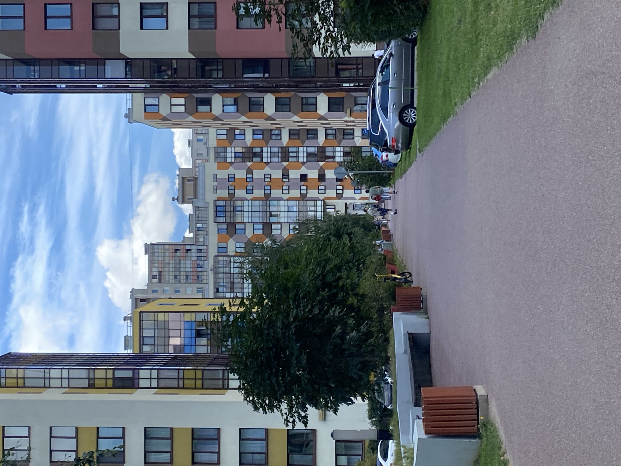 In Kudrovo, Russia, a residential area is depicted under a partly cloudy sky. The scene showcases a pathway leading into the distance, flanked by modern apartment buildings with multi-colored facades. On the left, a yellow and white building features numerous windows. To its right, a taller building with a mix of yellow and brown geometric patterns dominates the middle ground. On the far right, another apartment building exhibits a palette of pink, brown, and white blocks. Green trees and bushes line the pathway and the base of the buildings. Parked cars, including a silver sedan, are visible on the grass beside the road. A few individuals are strolling along the path, and a yellow scooter is parked nearby. Concrete benches and planters with wooden slats are situated along the pathway. The overall impression is that of a well-maintained urban living space.