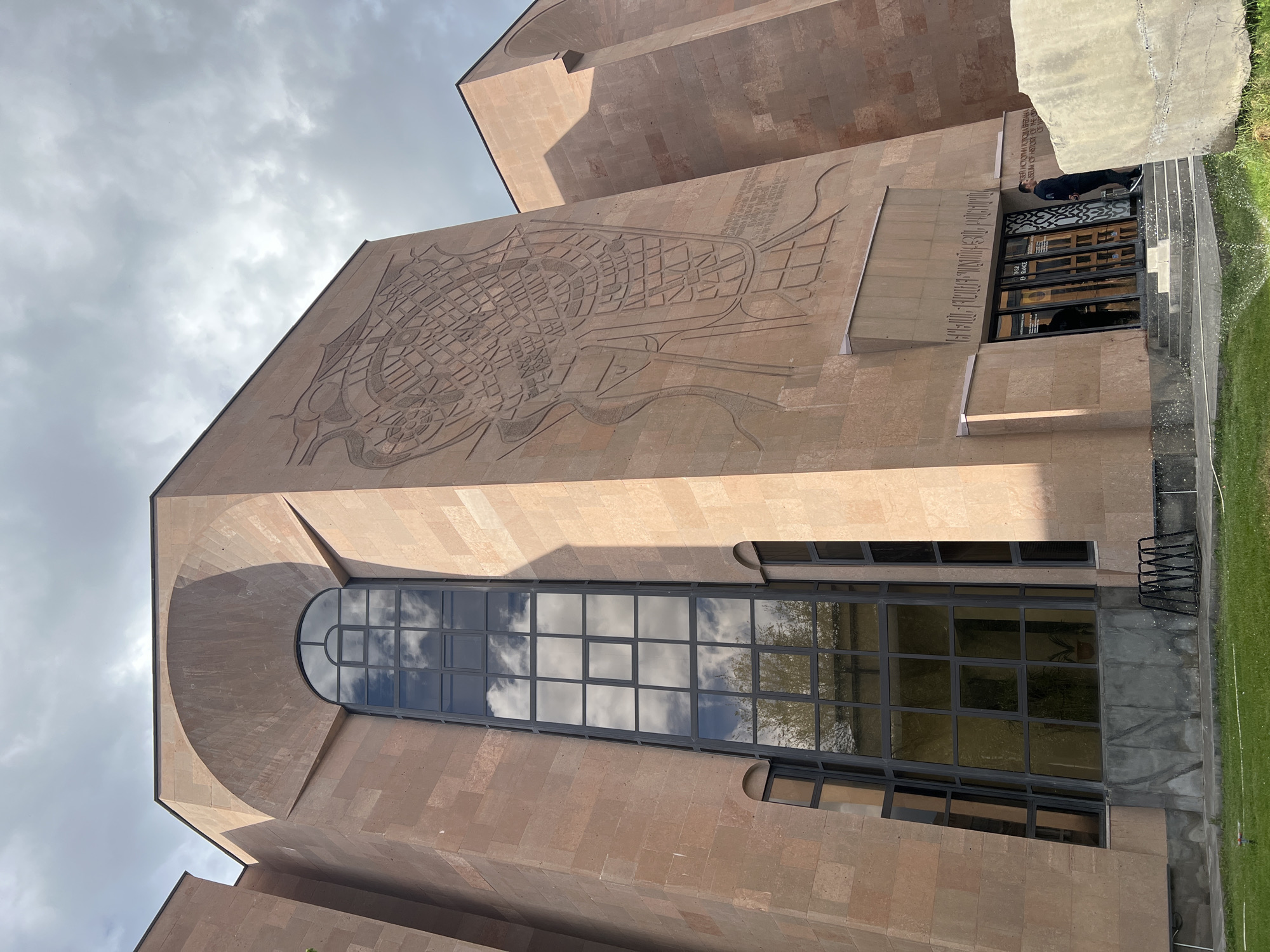 This media file presents a low-angle view of a monumental building, likely a museum or significant institution, under an overcast sky. The structure is constructed from light brown or peach-colored stone, exhibiting a modern architectural style with substantial, angular forms.
A prominent feature of the facade is a large, intricate bas-relief engraving that resembles a stylized city map or an ancient urban plan, complete with street-like patterns and curvilinear elements. Below this engraving, near the entrance, significant text is visible in both Armenian script and English: "ՀԱՅԱՍՏԱՆԻ ՊԱՏՄՈՒԹՅԱՆ ԹԱՆԳԱՐԱՆ" and "MUSEUM OF HISTORY OF ARMENIA," indicating the building's identity as the History Museum of Armenia.
The building features several windows. A large, arched window on the upper left section of the facade reflects the cloudy sky. Further down, a long, rectangular window stretches across the mid-section, also mirroring the sky and some nearby trees. The main entrance area consists of large glass doors, which have signage displaying "ԲԱՑ Է" and "OPEN," indicating the museum is currently open. A person, possibly a security guard or staff member, is visible standing near the entrance steps, dressed in dark attire.
In the foreground, a strip of green grass borders the building, with an active sprinkler system spraying water, suggesting maintenance of the grounds. The overall scene is captured during daytime, characterized by diffuse light due to the cloudy weather. The location is Yerevan, Armenia, as indicated by the context.