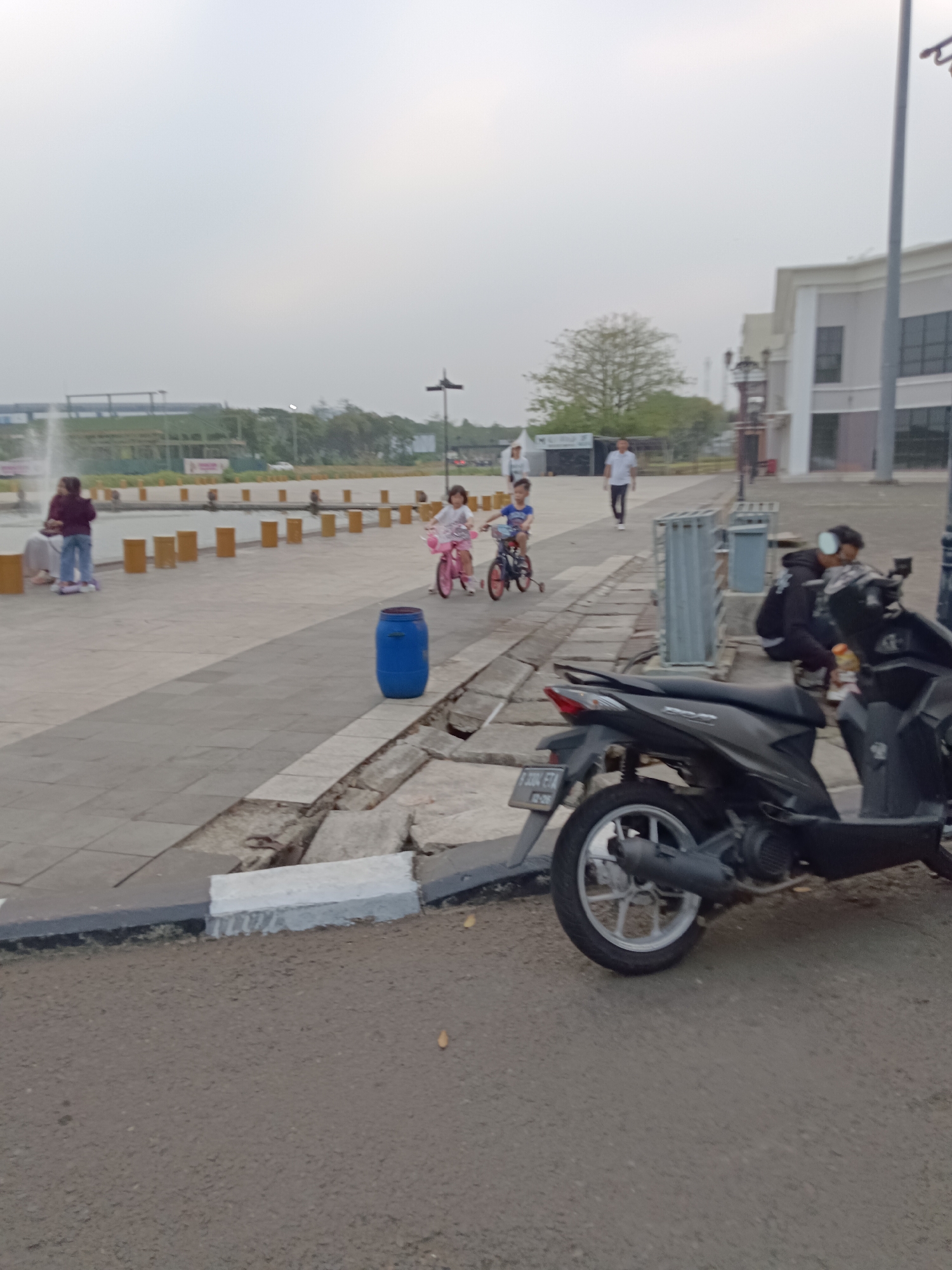 The image depicts an outdoor scene in what appears to be a public space in Jayamukti, Indonesia. In the foreground, a dark-colored scooter is parked on an asphalt surface. The scooter has a license plate that is partially visible. Behind the scooter, a concrete walkway is visible, marked with a white and dark grey curb. Further back, several people are present. Two children are riding bicycles on the walkway. One child, wearing pink, is on a pink bicycle, while another child, wearing blue, is on a blue bicycle. A blue barrel is positioned on the walkway near the children. To the left, a person in a maroon jacket and blue jeans is standing near a fountain. Other people are seen walking in the background. Beyond the walkway, there is a body of water with posts extending from it, and a building with a white facade and windows is situated in the background. Trees and other structures are also visible in the distance. The sky is overcast.