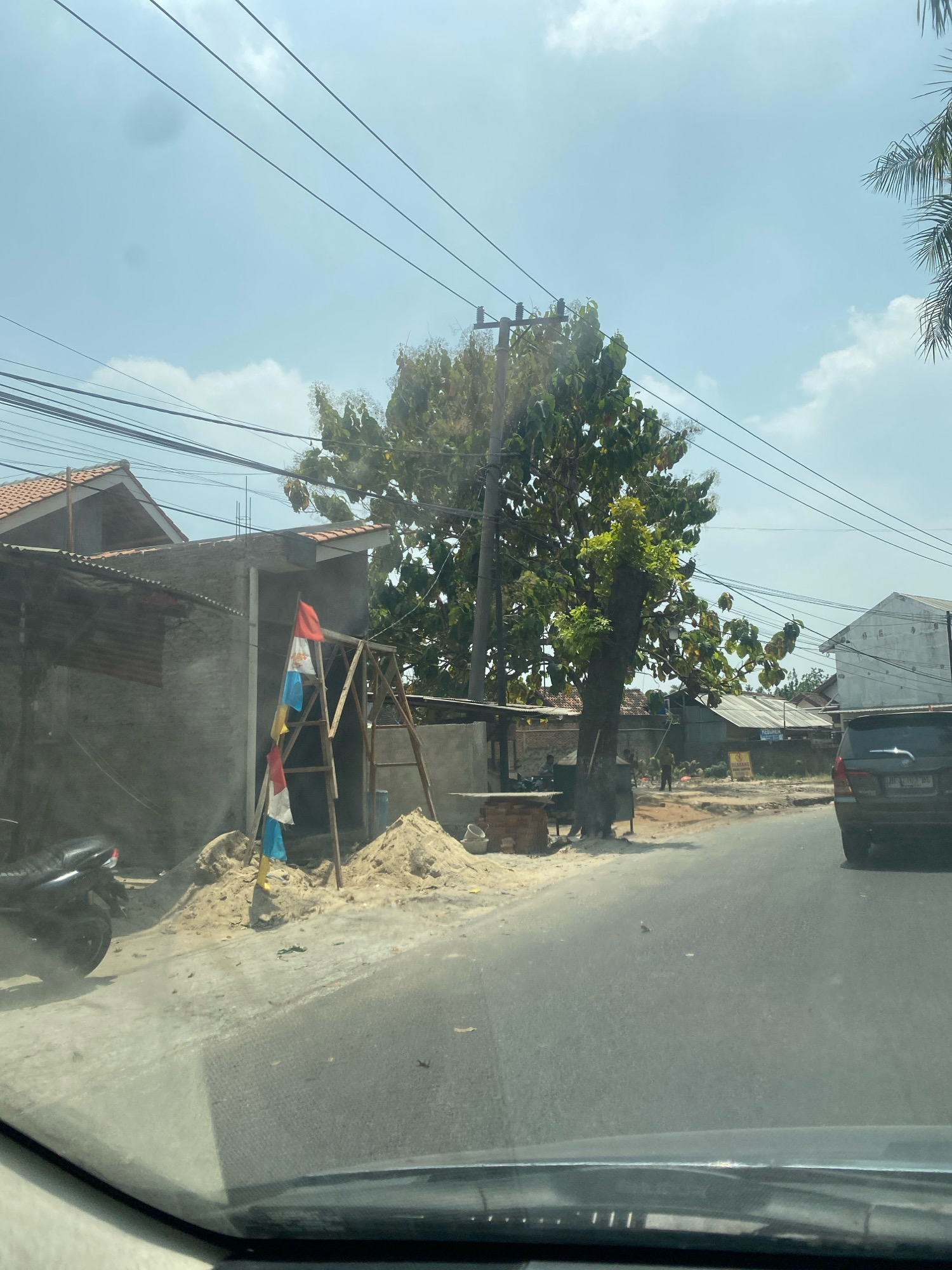 The image shows a roadside construction scene in Bandar Lampung, Indonesia. A partially constructed building with a concrete exterior and a tiled roof is visible on the left. A wooden scaffolding structure stands in front of the building, adorned with three flags: one red and white, one blue and white, and another red and white. Piles of sand and building materials are scattered around the construction site. A black motorcycle is parked to the left of the scaffolding. A large tree with dense foliage stands behind the scaffolding, partially obscuring a utility pole and overhead wires. In the background, other houses and buildings can be seen, along with a road curving to the right. A dark-colored SUV is driving away on the right side of the road. The sky is cloudy and bright.