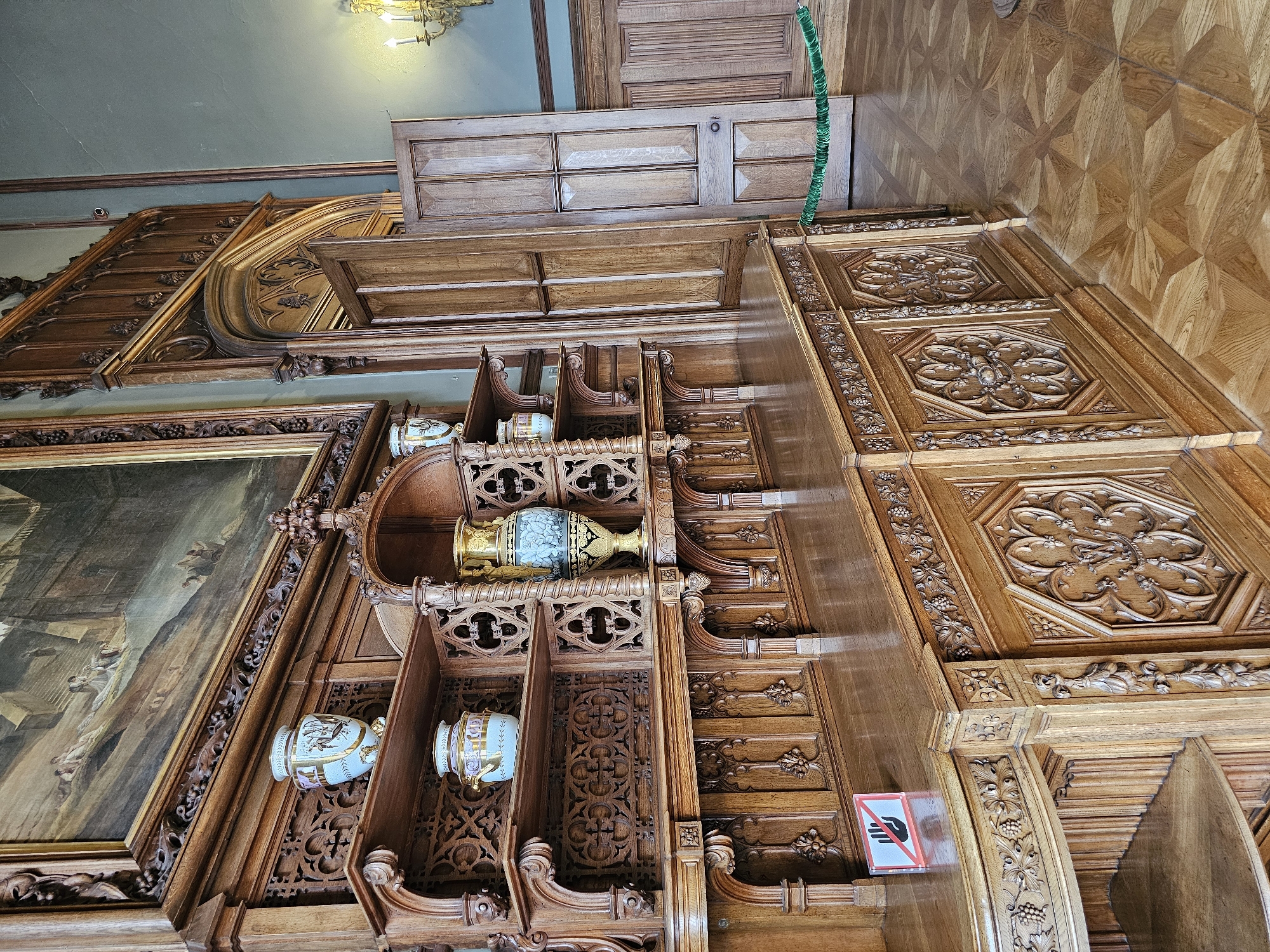 Captured from a low-angle perspective, the image showcases a segment of an ornately designed interior, presumably within the city of Alupkinskiy gorodskoy sovet, Ukraine. Predominantly crafted from wood, the scene features a richly carved wall structure, adorned with intricate patterns and floral motifs. The wall incorporates shelves that house decorative vases. A large painting with a gilded frame is visible to the left. Above, a wooden ceiling panel and a glimpse of a patterned wooden floor add to the scene's opulence. A 'Do Not Touch' sign is affixed to the wooden structure. The upper area of the photograph captures elements of the building's architecture, including a window and a lighting fixture.