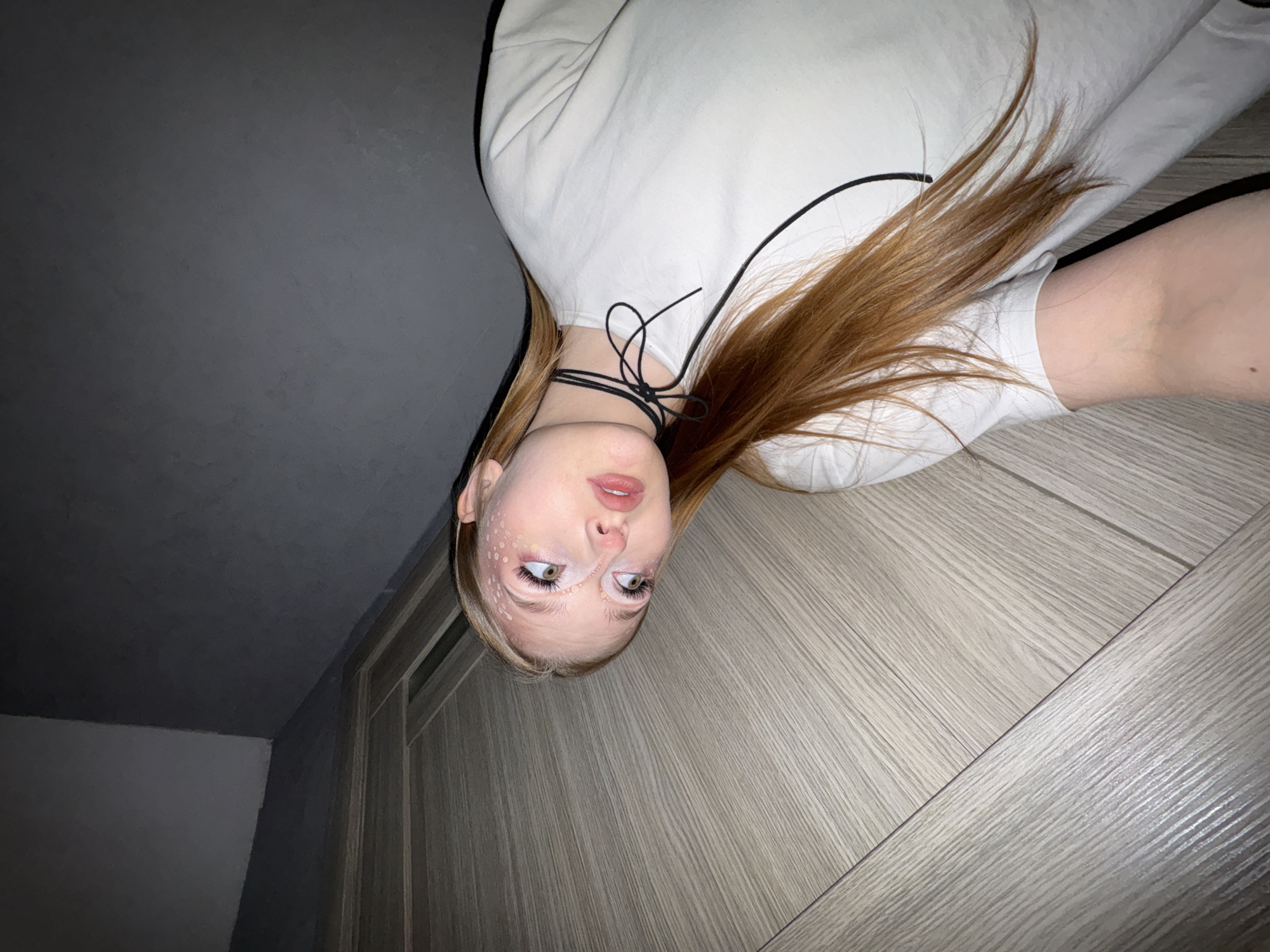 The image displays a young person, likely female, with fair skin and long, straight reddish-brown hair, positioned as if lying down or with the camera inverted. Her head is towards the bottom of the frame, gazing upwards. She wears a simple white long-sleeved top or sweatshirt, accented with thin black trim along the edges. A black choker necklace is tied around her neck in a delicate bow.
Her face is the central focus, featuring unique makeup: small white dots are artfully applied around her eyes and extend along the bridge of her nose. Her lips are slightly parted, adorned with a subtle light pink lipstick. Her light-colored eyes are wide open, conveying a sense of curiosity or mild surprise as she looks towards the viewer.
The background is divided diagonally into two distinct sections. On the upper right side (relative to her body), there's a light greyish-brown surface with a visible wood grain texture, resembling a laminate floor or a wooden door. On the lower left side, the background transitions to a dark grey, appearing to be a wall or ceiling, with a subtle corner visible, suggesting an indoor architectural detail. The overall lighting is soft and somewhat dim, contributing to a calm atmosphere.
The person is clearly posing for a photograph, with the distinctive makeup suggesting a creative or themed portrait session. The setting is an indoor space, likely a room within a residence. This photograph was taken in Chaikovsky, Russia. There is no visible text in the image.