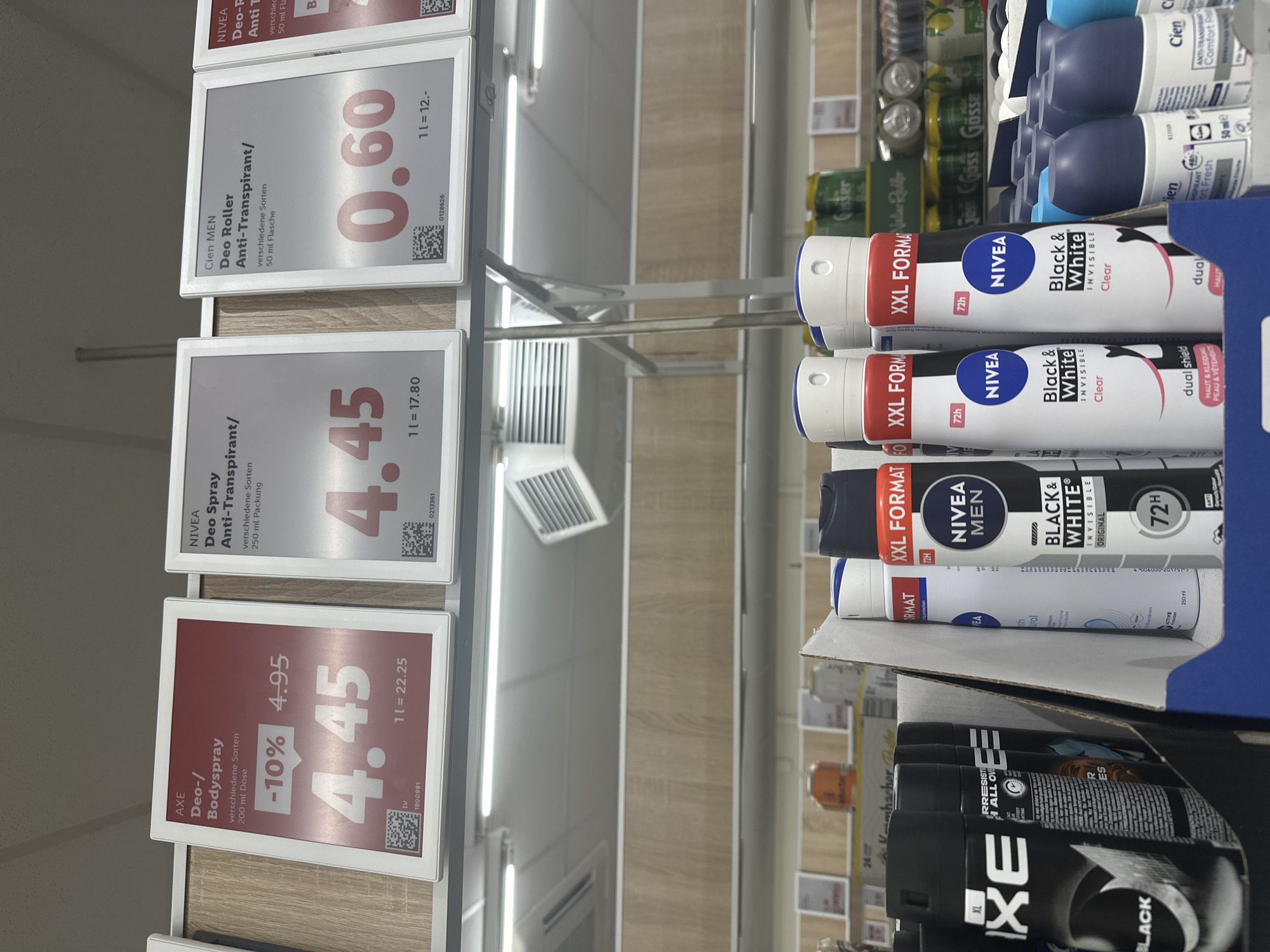 The image depicts a retail shelf display in a German supermarket, likely in Freiburg im Breisgau, Germany, showcasing various deodorant products. In the foreground, several aerosol deodorants are visible, including "AXE" and "NIVEA MEN Black & White." The shelf is also stocked with roll-on deodorants from "Cien." Above the products, illuminated price tags are displayed for "AXE Deo-/Bodyspray," "NIVEA Deo Spray Anti-Transpirant," and "Cien MEN Deo Roller Anti-Transpirant." The price tags indicate discounts and per-liter pricing. The overall environment appears to be a well-lit store aisle with typical retail shelving and overhead lighting.