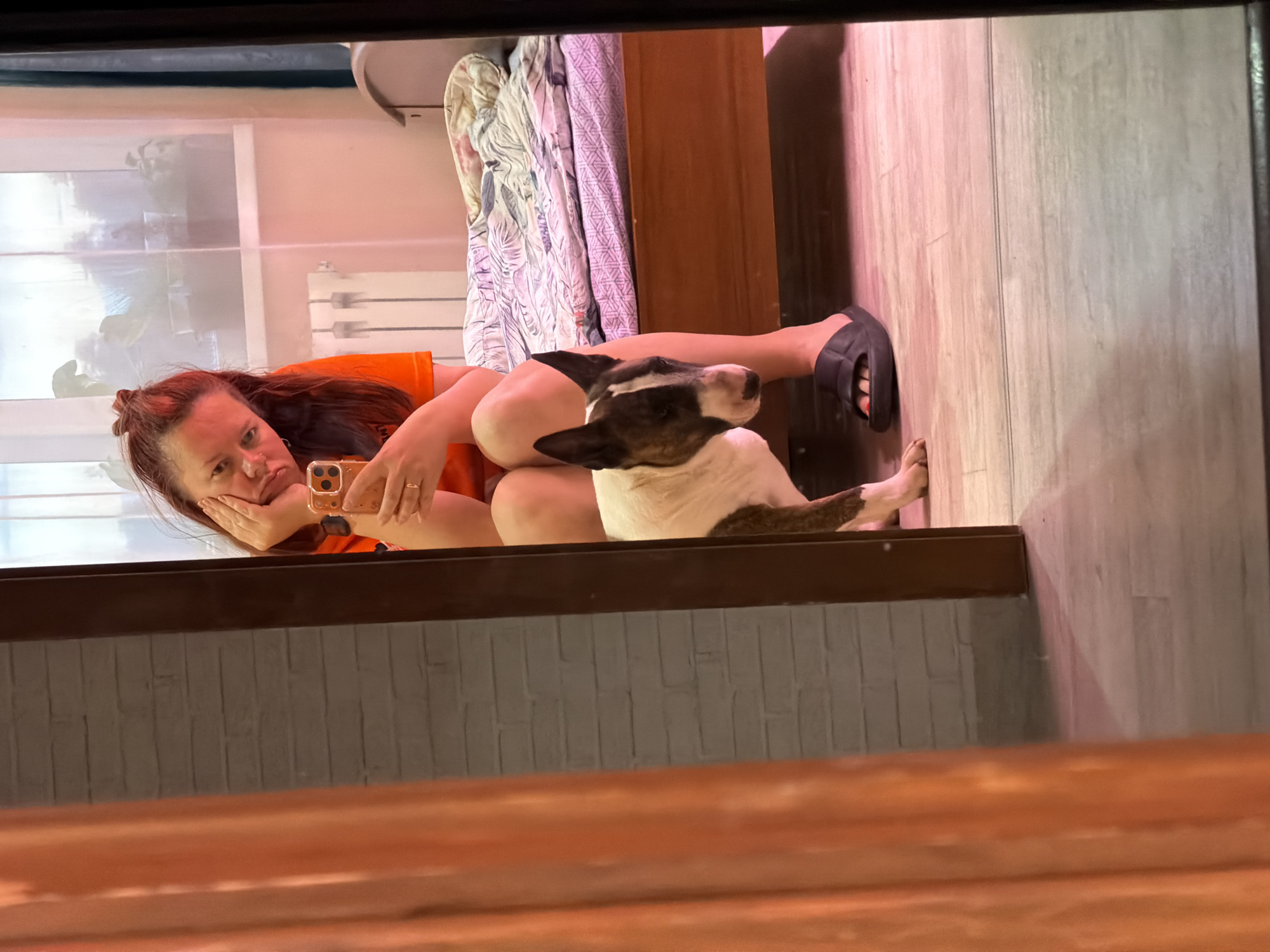 The image, taken in Chaikovsky, Russia, shows a woman and a dog in a relaxed indoor setting, likely reflected in a mirror or glass surface. The perspective is unusual, with the subjects appearing upside down as if viewed in reflection, or the image itself is rotated.
The main subjects are a woman with reddish-brown hair, partially tied up, and a Bull Terrier dog. The woman is wearing an orange top and dark sandals. She has a ring on her left hand. She is holding a gold-colored smartphone, seemingly taking a selfie, and rests her chin on her hand, displaying a slightly pouty or thoughtful expression. Her other arm is bent. The dog, which is white with dark brown or black patches on its head and body, lies beside her, looking upwards and slightly away from the camera.
The background reveals a room with light-colored wooden or laminate flooring. A window with a white radiator below it is visible, suggesting daylight, although the indoor lighting has a warm, possibly pinkish, hue. Also visible are parts of a bed with patterned bedding and what appears to be a wooden closet or furniture. A dark wooden frame or ledge runs across the foreground, indicating the edge of the reflective surface. There is no discernible text visible in the image. The overall mood conveyed is casual and relaxed, perhaps a moment of downtime with her pet.