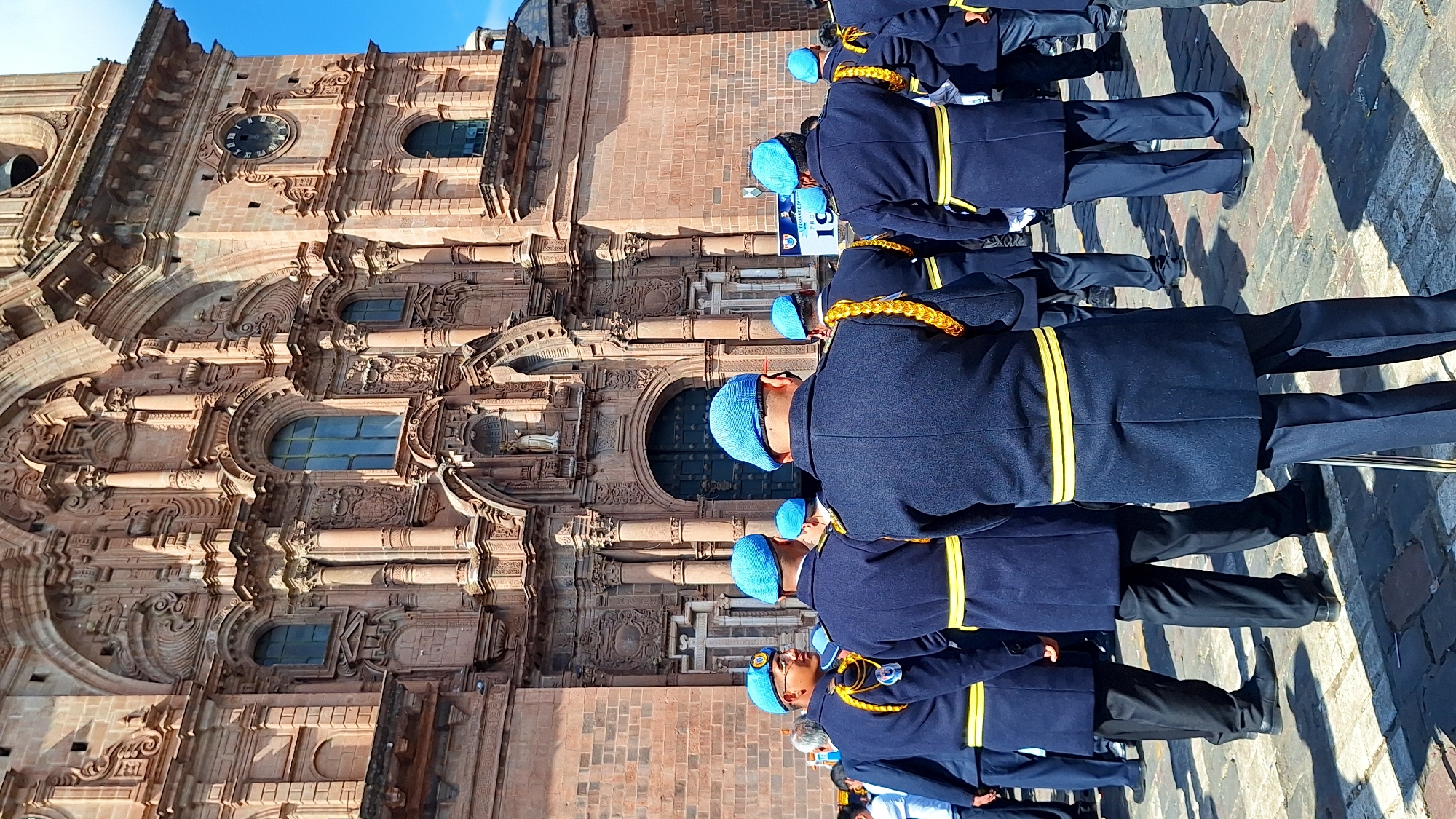 The image shows a group of individuals in dark blue uniforms with yellow sashes and light blue berets, standing on a cobblestone plaza in front of a large, ornate building. The building appears to be a church or cathedral, constructed of reddish-brown stone with intricate carvings, arched windows, and a prominent clock tower. The architectural style suggests it is likely the Cusco Cathedral, located in the Plaza de Armas in Cusco, Peru. Some individuals are facing away from the camera, while others are looking to the side. The weather appears to be sunny, with clear blue skies visible above the building. A banner with Spanish text and the number "19" is partially visible to the right of the frame. The individuals are standing in a formation, suggesting a parade or ceremony.