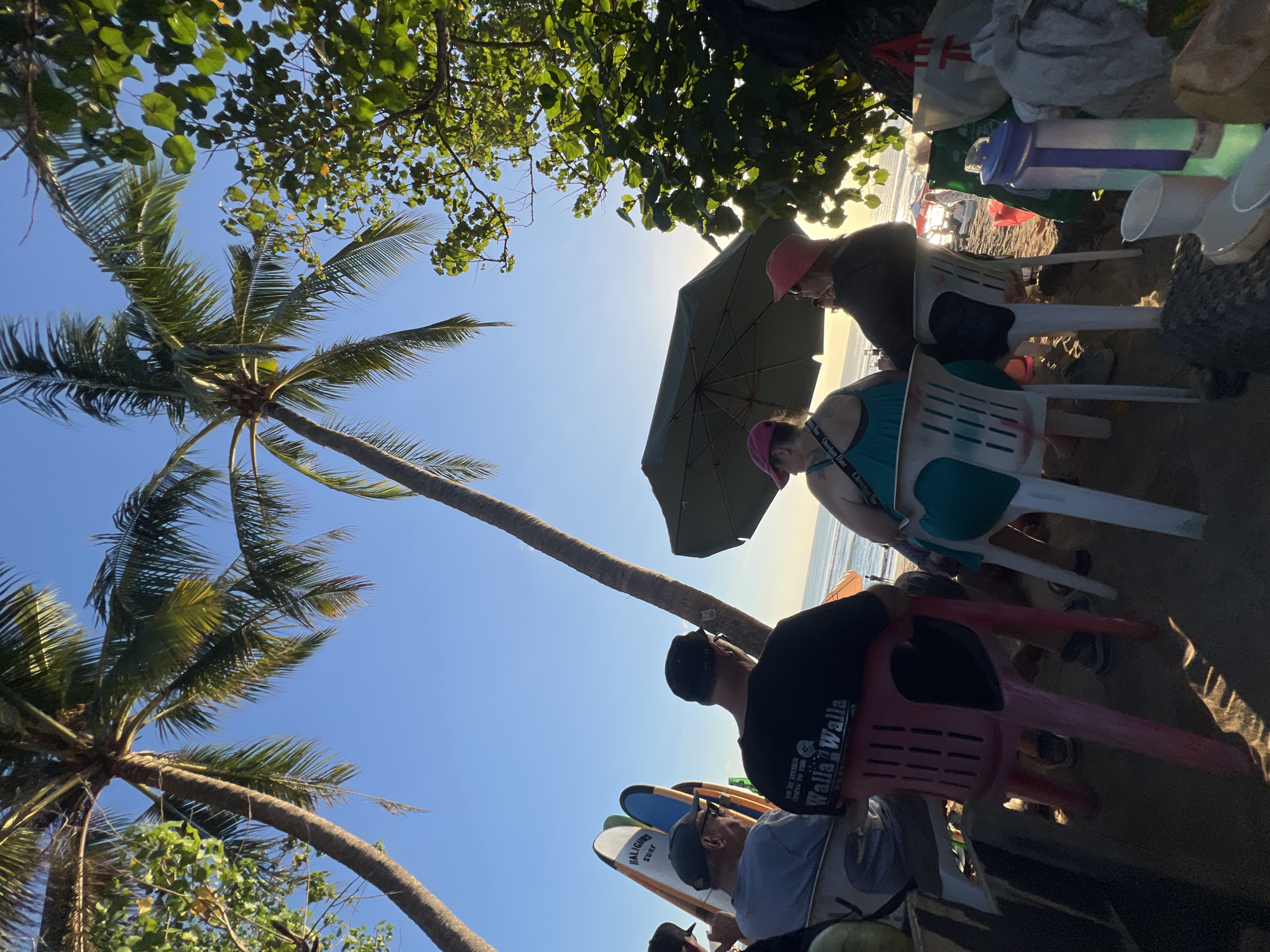 The image depicts a beach scene in Kuta, Indonesia, at sunset. Several people are seated on plastic chairs on the sandy beach, with palm trees and lush green foliage framing the view. The sun is low in the sky, casting a warm glow and long shadows.
In the foreground, a group of individuals are relaxing. One person in a black t-shirt with "Walla Walla" printed on the back is visible. Next to them, another person wears a turquoise top and a pink hat. Further to the left, two men are seated, with surfboards leaning against a wall behind them. The surfboards are branded "Baliguns Surf".
An open green umbrella provides shade for some of the seated individuals. The ocean is visible in the background, with gentle waves lapping the shore. The sky is clear and blue, transitioning to a warmer hue near the horizon. Various bags and water bottles are scattered around the seating area, suggesting a casual gathering. The overall atmosphere is relaxed and serene, capturing a moment of leisure on the Kuta beach.