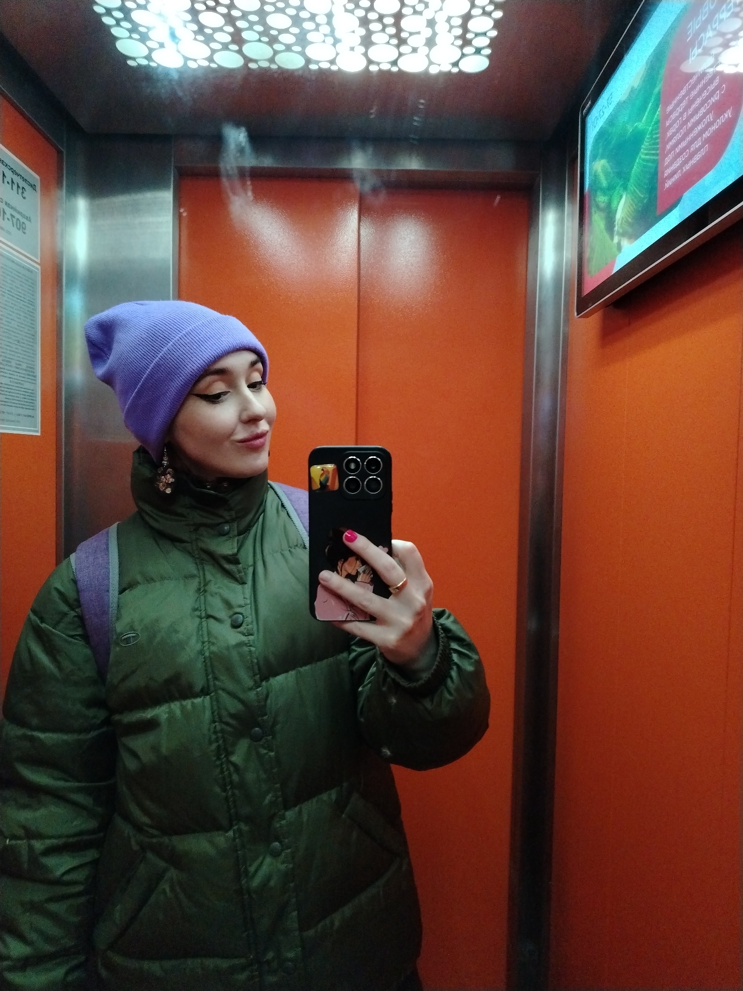 The image is a selfie taken in an elevator. The subject is a woman with a light complexion, wearing a lilac beanie, a green puffer jacket, and earrings. She is holding a black smartphone with a custom case, presumably taking the selfie. The elevator's interior features orange walls, a silver metal frame, and a digital display screen in the upper right corner showing text in a Cyrillic alphabet. There is also an information panel visible on the left wall of the elevator. The lighting appears to be provided by a patterned ceiling fixture. The elevator seems to be located in a building in Izhevsk, Russia.