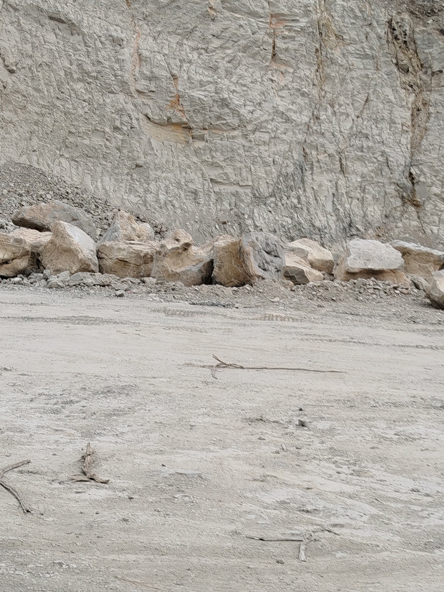 This image depicts a quarry with a sheer rock face in the background, predominantly light gray in color with visible strata and some reddish-brown mineral deposits. In the mid-ground, a collection of large, rounded rocks lies at the base of the rock face, interspersed with smaller gravel. A dirt track or road occupies the foreground, showing tire marks and scattered debris, including pieces of dried vegetation. The scene is likely located in Kemangguhan, Indonesia, given the context provided. There are no people or active operations visible in the image. The overall impression is one of a raw, natural landscape modified by quarrying activities.