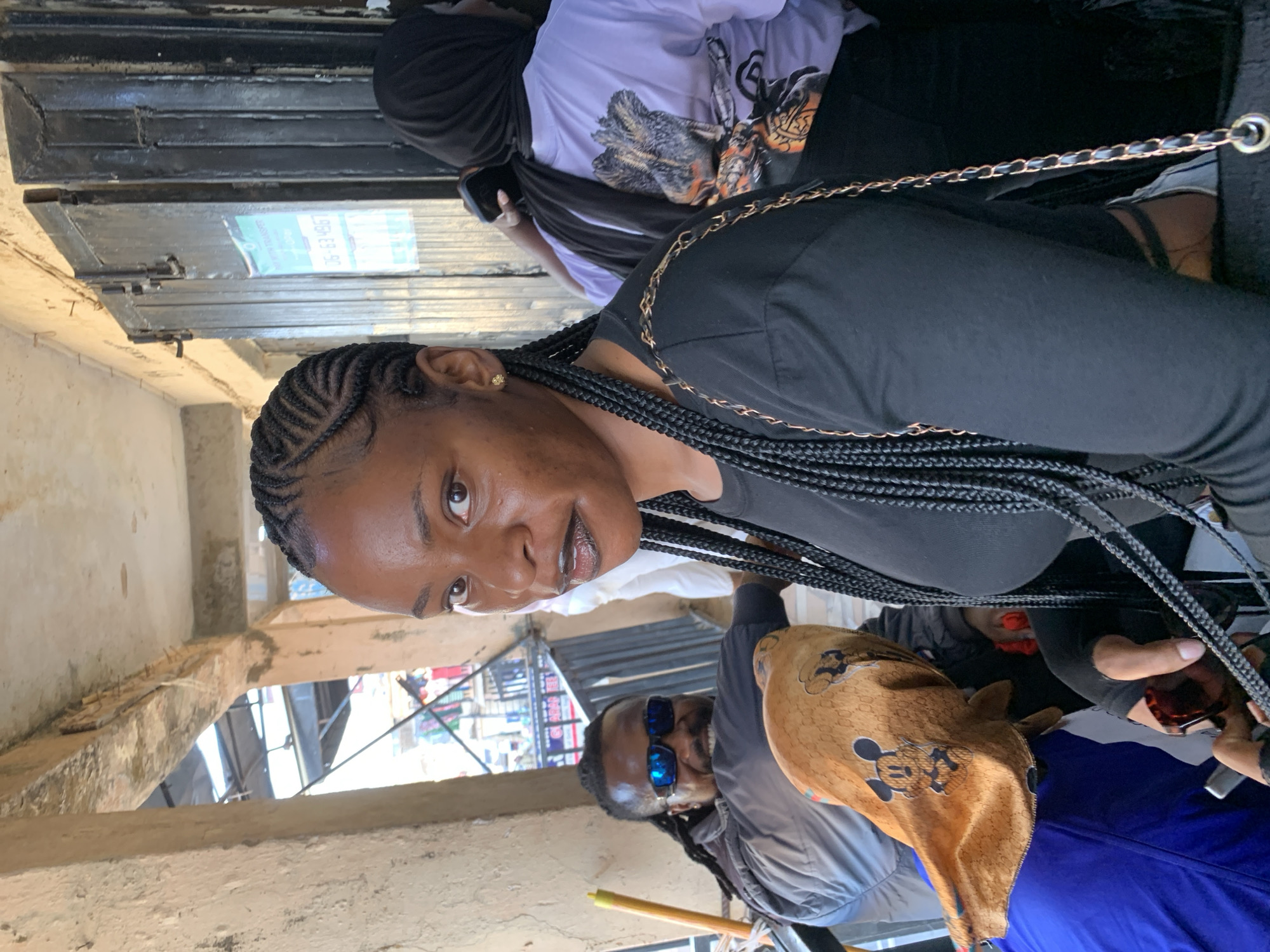 The image depicts a group of people in an urban setting in Jos, Nigeria.
The main subject is a Black woman with braided hair, wearing a black top. She is the focal point of the image. Behind her, another person is visible, wearing a light purple shirt and a black head covering.
Further down, two other individuals are present. One wears blue sunglasses and a light grey jacket. The other person is wearing a Mickey Mouse themed patterned material.
The setting appears to be a building, likely a market with metal bars and concrete walls. There are signs of construction or exposed framework. The background shows a glimpse of the city.
The scene is brightly lit, indicating daytime.