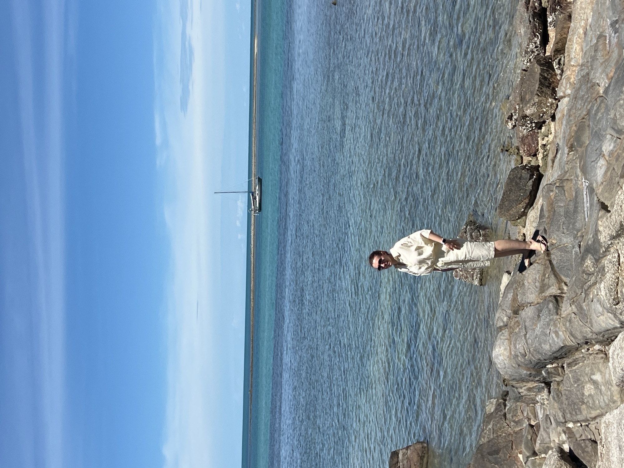 A full-shot photograph depicts a woman standing on a rocky shoreline. The woman, positioned slightly right of center, faces the camera and smiles. She has short, dark hair and wears sunglasses. Her attire consists of a light-colored, collared button-up shirt and matching shorts. She is also wearing sandals. The shoreline is composed of large, grey rocks and pebbles, with a scattering of smaller rocks and some vegetation visible in the lower right corner.
The ocean stretches out behind her, with clear, blue-green water. A distant sandbar or shallow area is visible as a lighter brown strip parallel to the horizon. Further out, a single sailboat with a tall mast is anchored on the water. The sky above is a vibrant blue with thin, wispy white clouds. The lighting suggests it is a sunny day. The scene appears to be a coastal location, likely in Baan Hua Thanon, Thailand, given the clear tropical waters and shoreline features.