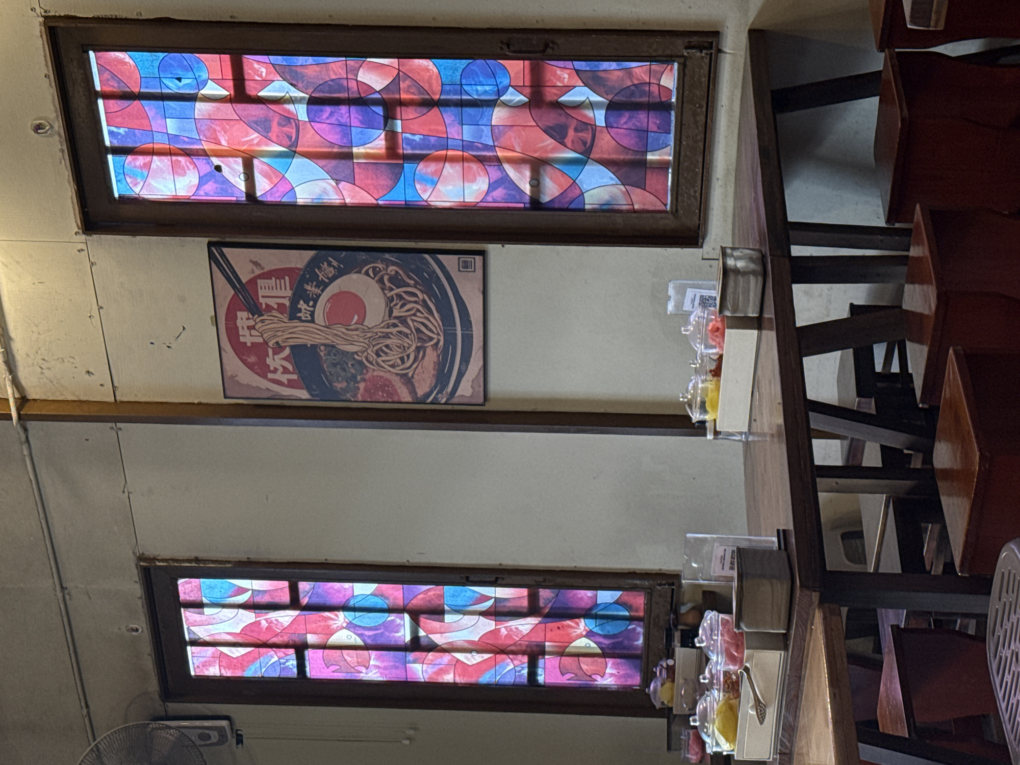 The image depicts the interior of a restaurant in Lâm Viên - Đà Lạt, Vietnam. Two tall, narrow stained-glass windows with abstract patterns in shades of red, purple, and blue flank a framed poster of a ramen bowl. A dark wooden table with several chairs and stools is set in front of the windows. On the table are small glass containers with condiments and a silver serving spoon. The walls are a neutral color, and the ceiling is visible at the top of the image. A vintage fan is mounted on the left wall. The overall ambiance suggests a cozy, possibly retro dining establishment.