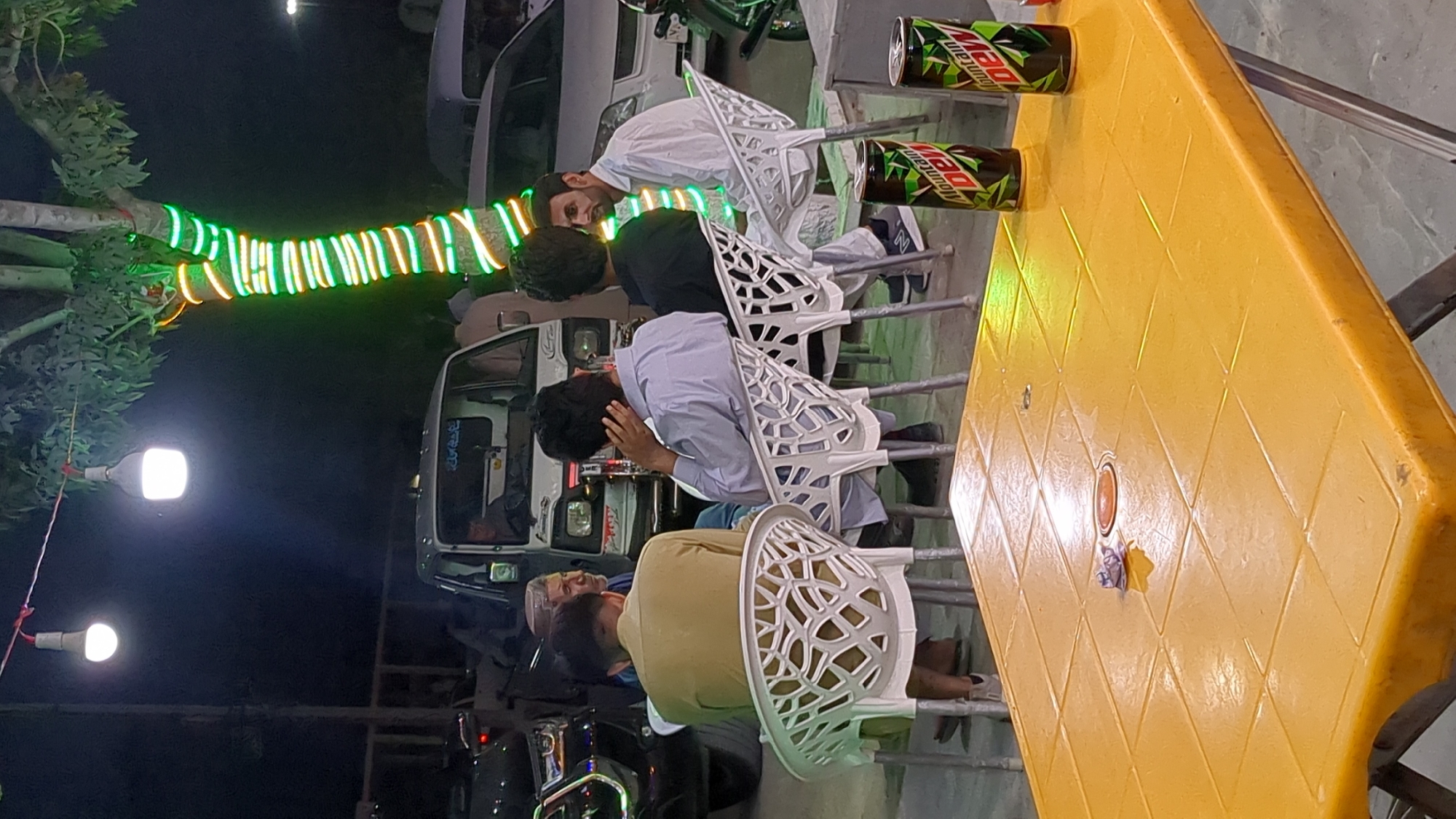 The image depicts a nighttime scene at an outdoor eatery in Quetta City Tehsil, Pakistan. Five people are seated on white plastic chairs around a yellow plastic table. Two cans of Mountain Dew are on the table's edge. A tree is decorated with green and orange lights. A minivan is parked nearby. A white light source is visible in the upper left.