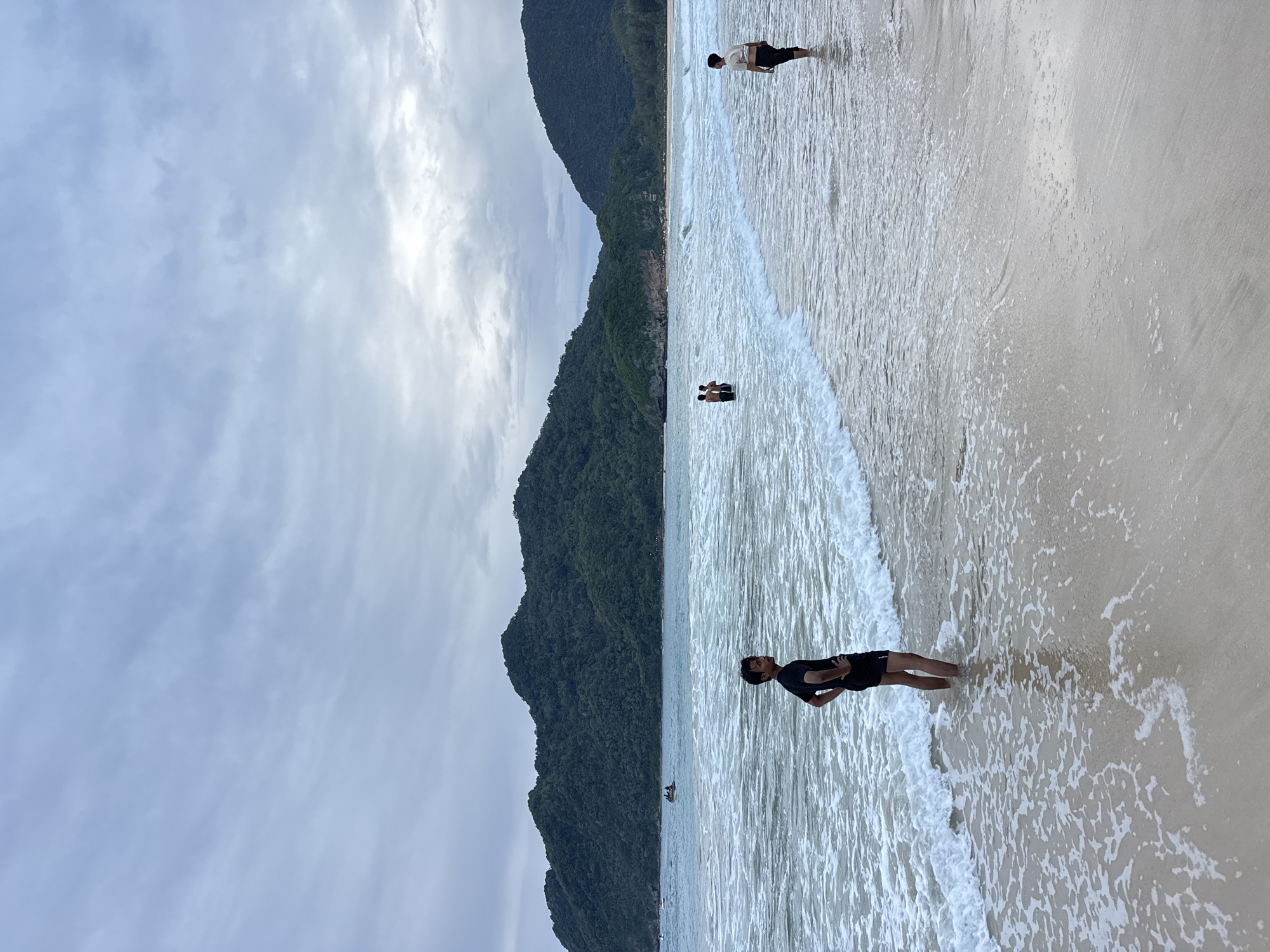 This image depicts a beach scene in Meunasahbale, Indonesia. The sky is overcast with a light blue hue. In the foreground, a man stands in shallow water, facing to the right with his hands on his hips. His attire consists of a black t-shirt and black shorts. Further out in the water, two people are visible, appearing to be standing or wading. To the right, another person stands in the water, facing towards the shore. The sea exhibits moderate waves with white foam. In the background, lush green, tree-covered hills form a scenic backdrop. The beach itself has a sandy surface.