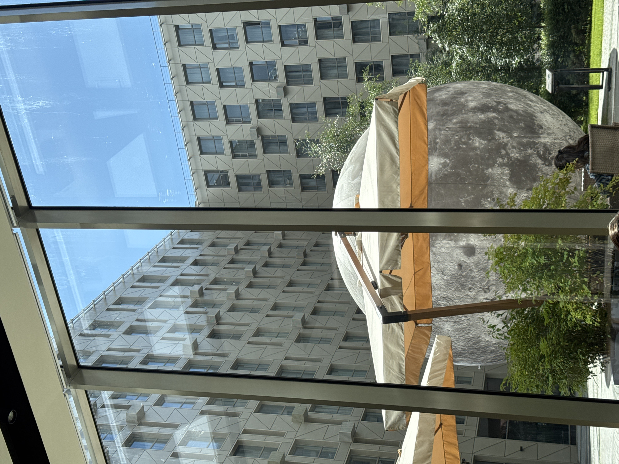 The image shows an outdoor courtyard viewed through a large window with vertical metal framing. In the foreground, there are two large, cream-colored umbrellas with orange trim, providing shade. Behind the umbrellas, a massive, spherical sculpture resembling the moon is prominently displayed. The moon sculpture has a textured surface with craters, typical of lunar depictions.
Surrounding the moon sculpture is lush green foliage, with bushes and trees. To the left and right of the moon sculpture, modern, multi-story buildings with numerous windows are visible. These buildings feature a repeating geometric pattern on their facades, and some have visible air conditioning units mounted on the exterior. The sky above is a clear, bright blue.
In the lower right corner of the image, a person is partially visible, sitting in a wicker chair, facing away from the camera. They have long, dark hair tied back. A small, dark-colored signpost stands on the grass to the right of the moon sculpture. The scene appears to be in a public space, possibly a hotel courtyard or a plaza.
This image was taken in Saint Petersburg, Russia, specifically in the courtyard of the Hotel Indigo St. Petersburg - Tchaikovskogo. The moon sculpture is a permanent art installation within the hotel's courtyard.