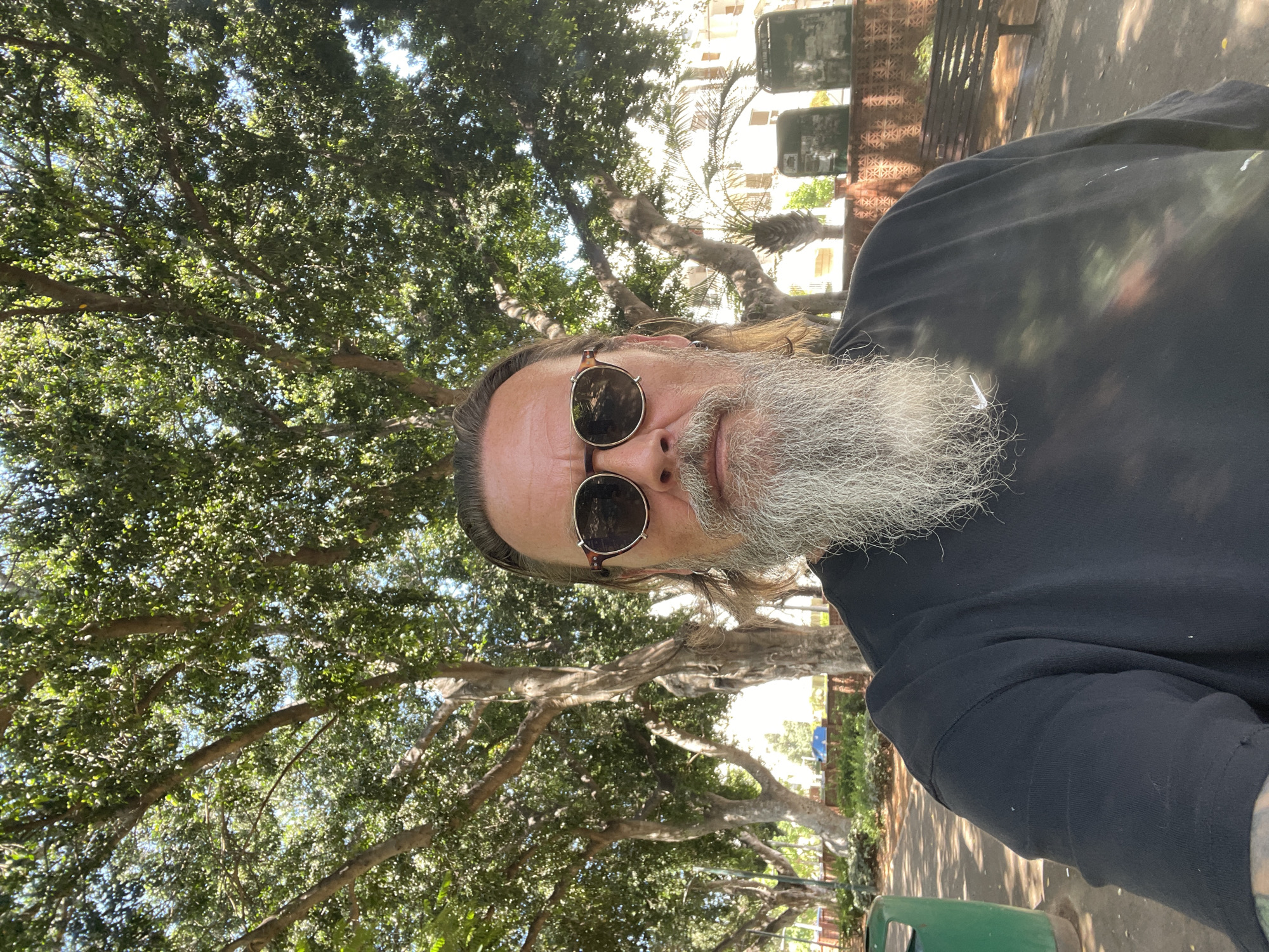 A medium shot selfie shows a man with a long grey beard and sunglasses standing outdoors under large trees. He is wearing a black t-shirt. The background includes more trees, some buildings, a trash can, and a bench. The scene appears to be in a park in Netanya, Israel.