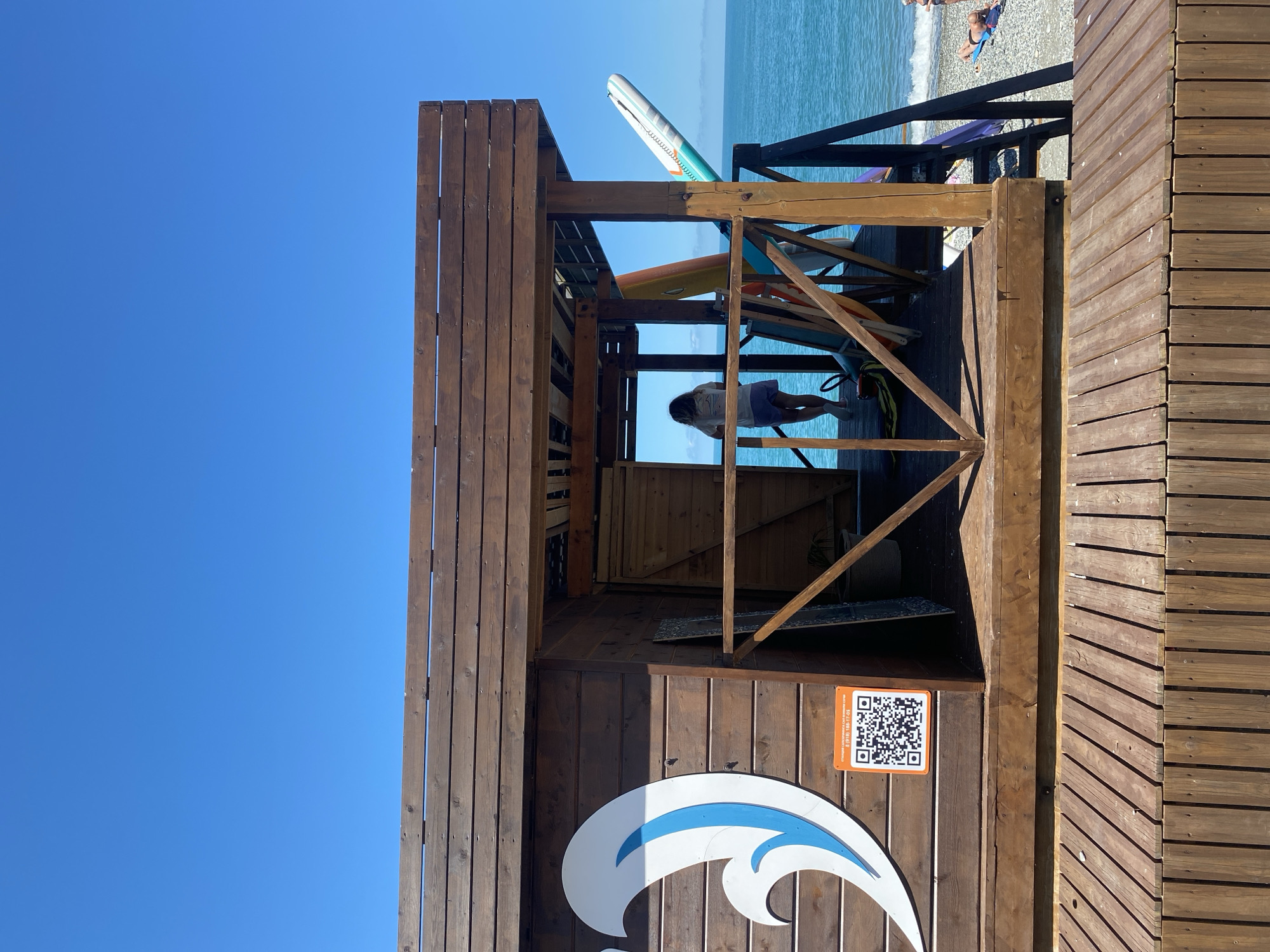 On a clear day in Sochi, Russia, a wooden structure, possibly a beach rental stand, stands on a pebble beach. The structure is made of horizontal wooden planks and has an open, covered area where a person is standing and looking out towards the sea. Inside the open area, surfboards and paddleboards are propped up against the back wall. To the left of the entrance, a large white wave logo with a blue outline is painted on the wooden wall, and below it is a QR code sticker. A wooden deck or walkway leads up to the structure. In the background, the blue sea meets the sky, and a few people are visible relaxing on the beach.