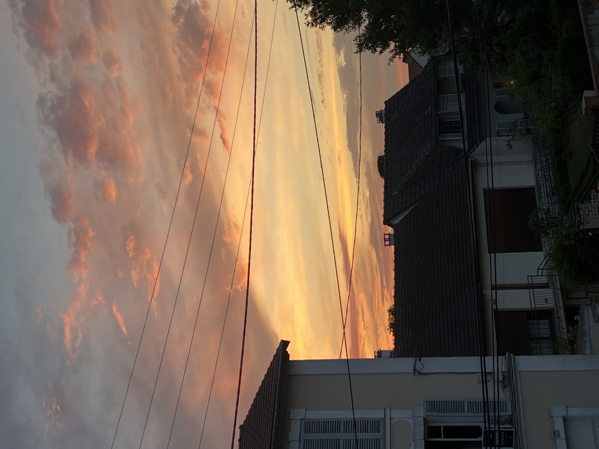 The image depicts a sunset over the city of Montfermeil, France, as viewed from a residential area. The sky is filled with dramatic clouds displaying a gradient of colors from pale grey to vibrant orange and pink, indicative of the setting sun. Several utility wires crisscross the frame in the foreground, obscuring parts of the sky and the rooftops of houses below.
In the lower left, a portion of a light-colored building with grey shutters on its windows is visible. Adjacent to it, several dark-tiled rooftops of residential homes are prominent. The architecture suggests a typical European suburban setting. The houses have chimneys and dormer windows. Some trees and greenery are present between and behind the houses, with dense foliage visible in the lower right quadrant. The overall scene captures a serene, yet visually striking, end-of-day atmosphere in Montfermeil. There are no people visible in the image.
