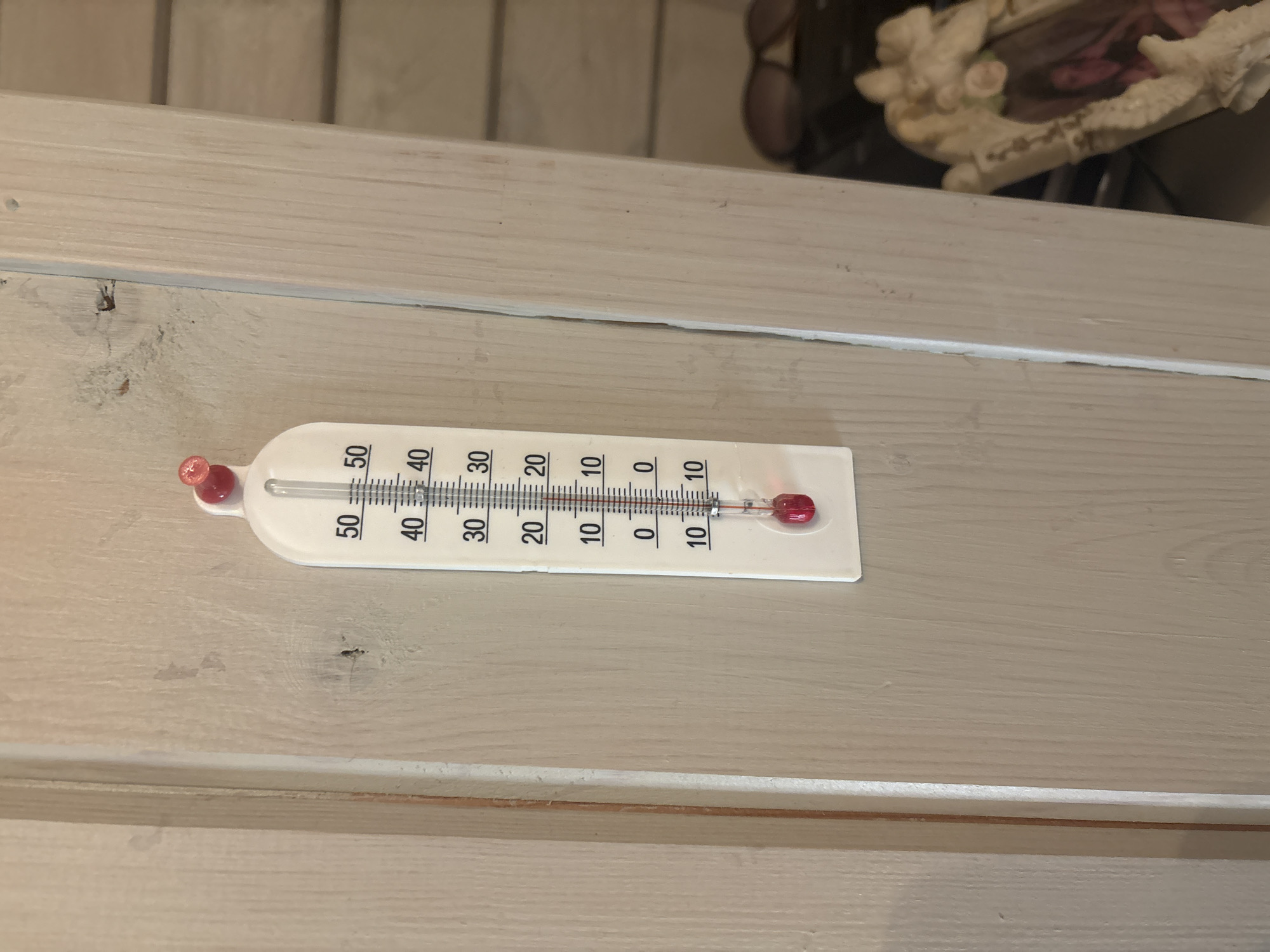A thermometer is affixed to a light-colored wooden surface, likely a door or wall. The thermometer, white with black markings, displays temperatures in Celsius ranging from 10 to 50 degrees, with increments of 10 clearly labeled. The red liquid within the thermometer indicates a temperature below 10 degrees Celsius, as it has not reached the 10-degree mark yet. The overall scene appears to be indoors or in a sheltered outdoor area, given the presence of a framed photograph in the background and the wooden construction. The context suggests a typical household setting in the city of Berezka Snt (Povarevo), Russia.