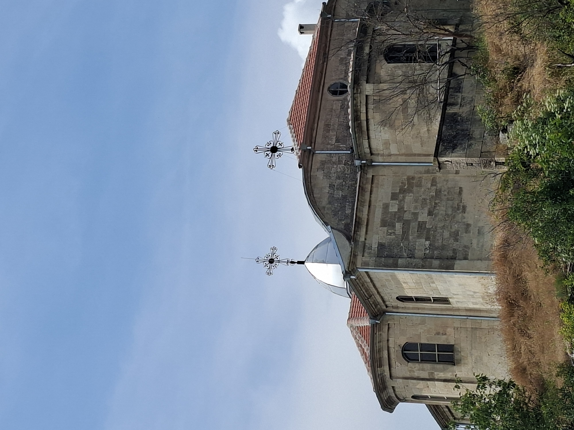 The image displays a stone church structure situated on a vegetated incline under a clear, light-blue sky with minimal scattered clouds. The scene is located in Basarbovo, Bulgaria.
The church building is constructed primarily of light-colored, cut stone blocks. Its architectural style features rounded sections and appears to be multi-faceted. The roofs are covered with reddish-orange terracotta tiles, and silver-colored rainwater downspouts are visible along the exterior walls. One prominent dome, silver and metallic in appearance, rises from the central part of the structure, topped with an ornate black metal cross. Another ornate black metal cross is positioned atop a tiled roof section to the right of the silver dome.
The building exhibits several windows: two tall, arched, multi-paned windows are visible on the left rounded section, and one circular window is present on the right-hand section of the building. A dark, possibly boarded-up or deeply shaded arched window is also visible on the far right. A small chimney stack protrudes from the tiled roof on the far right.
The immediate foreground and the sloped ground leading up to the church are covered with dry, tan-colored grasses and various green shrubs and bushes. A few bare tree branches are visible near the right side of the building. No human figures or discernible animal life are present in the image. The lighting suggests daytime conditions.