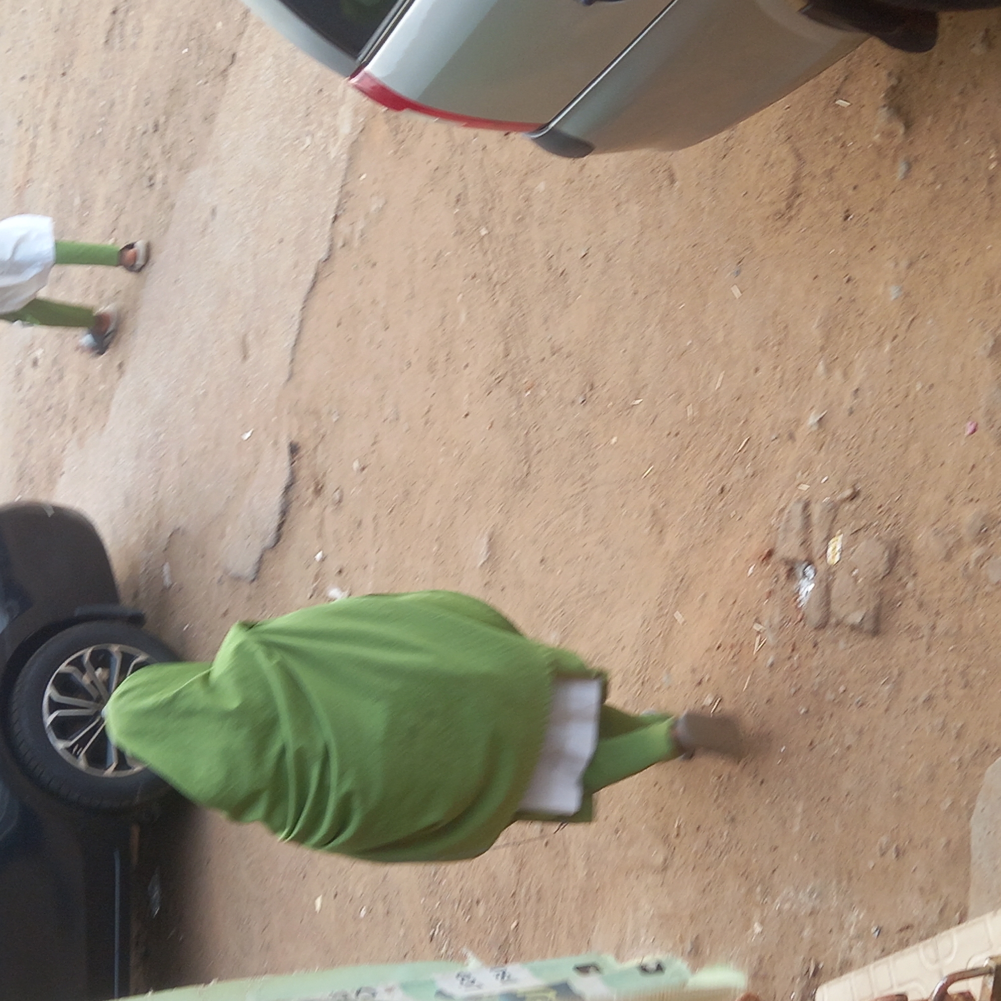 A top-down, ground-level shot shows a person walking on a dirt surface alongside vehicles, possibly a street in Dadi Riba, Nigeria. The person is wearing a green garment with visible white trim. Part of a silver car is visible in the top of the frame and a dark-colored car appears in the lower-left. Another person wearing a white top and green pants is visible in the upper-left. There is what appears to be a manhole cover in the center-right of the frame.