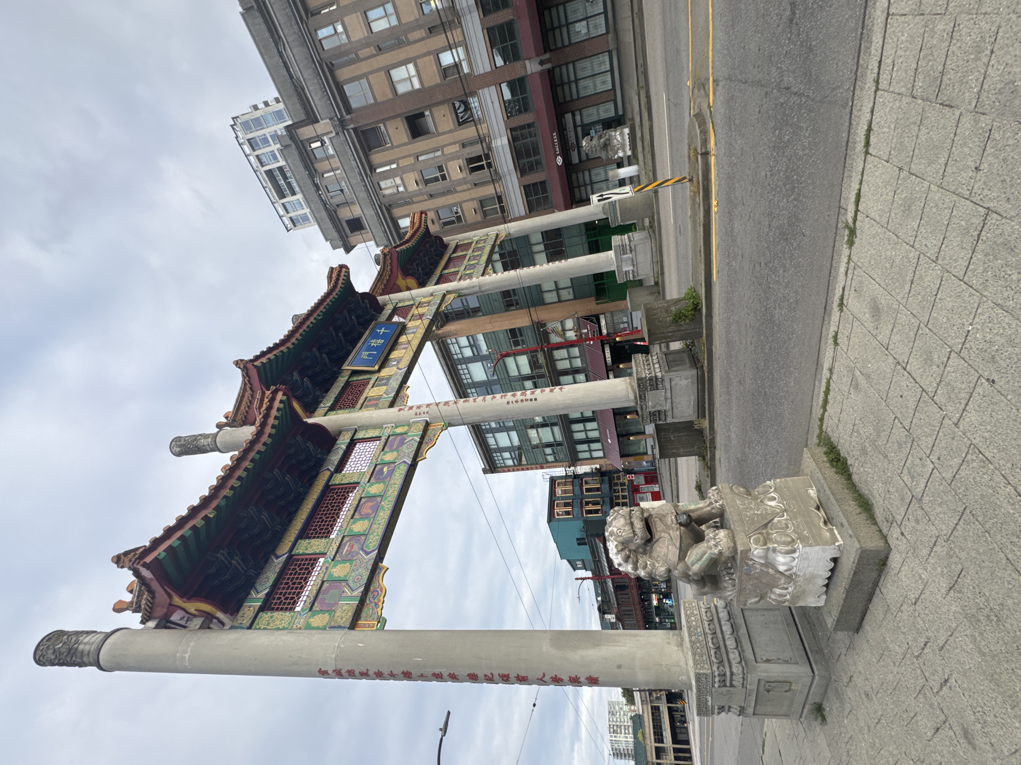 This is a full, wide-angle shot of the Chinatown Gate in Vancouver, Canada, on a cloudy day. The gate, a traditional Chinese archway with intricate colorful designs and Chinese characters, is the central subject. Two stone lion statues stand guard at the base of the gate. In the background, various multi-story buildings, some with traditional architecture and others more modern, line the street. The street itself is paved and appears to be a moderately busy urban thoroughfare. No people are visible in the image.
