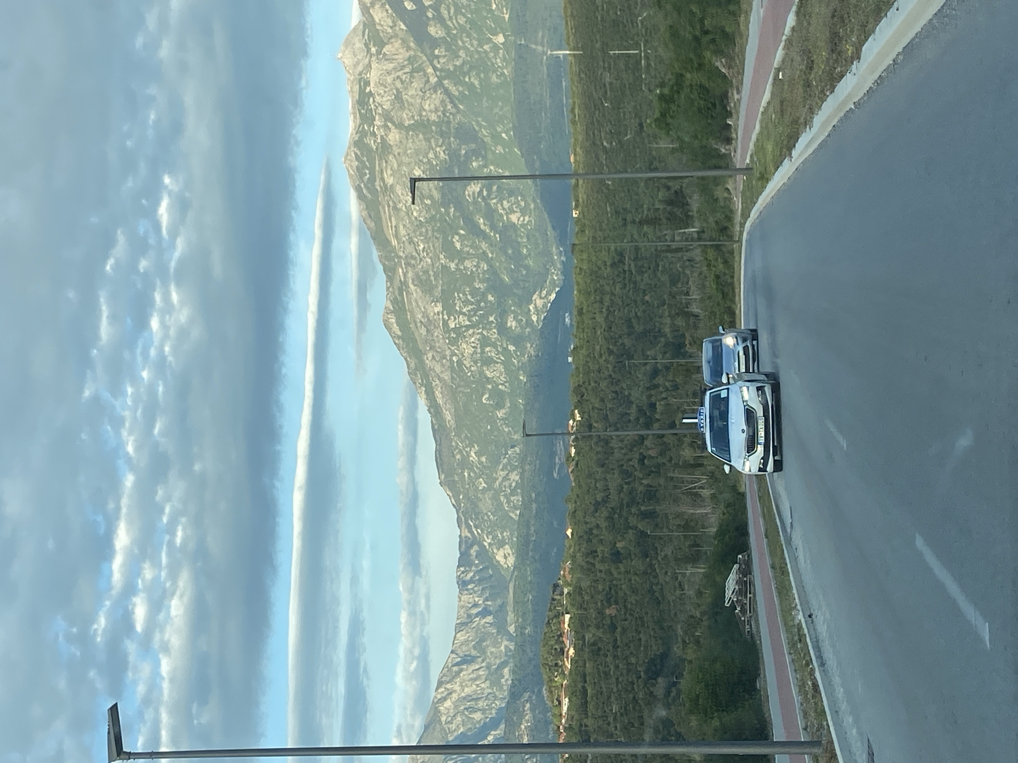 The image depicts a scenic road in Radovići, Montenegro, with rugged mountains and lush greenery under a partially cloudy sky. The perspective is slightly elevated, looking ahead and to the right, capturing a paved road that curves gently.
In the foreground, a white SUV or crossover, clearly marked with a "TAXI" sign on its roof, is driving on the road. Its license plate prominently displays "TV AX," indicating a registration from Tivat, a municipality in Montenegro. Behind it, a darker colored car is also on the road. A narrow red/brown path runs parallel to the main road on the right, separated by a low concrete barrier. Several tall, dark streetlights or utility poles line the roadside.
The midground features a dense, verdant forest or shrubland, covering undulating terrain that extends towards the mountains. The background is dominated by an impressive range of large, rocky mountains, their gray, textured surfaces showcasing various geological features.
The sky above is light blue with scattered clouds. A distinct, elongated cloud, possibly a contrail, stretches horizontally across the sky, while other clouds appear more diffuse and patchy. The natural lighting suggests either early morning or late afternoon, casting soft illumination across the landscape. The scene conveys a sense of travel through a beautiful, natural environment. No people are visible in the image.