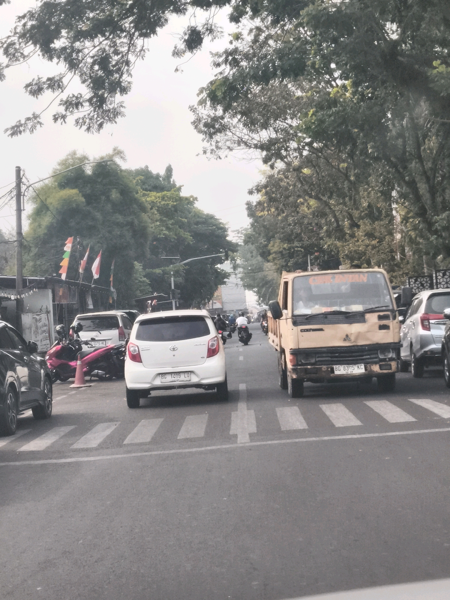 The image depicts a street scene in Palembang, Indonesia, likely during daylight. A yellow, older model Mitsubishi truck is prominent in the foreground, stopped at a crosswalk. The truck has "CIP AYAN" written in orange lettering on its windshield. It displays a license plate "BG 8305 KC".
To the right of the truck, a silver car is visible, and to the left, a white Toyota Agya with license plate "BG 1409 LU" is also stopped at the crosswalk. Behind the Toyota, a white SUV is parked. A pink motorcycle is parked next to the white SUV, with a traffic cone nearby.
In the background, trees line the street, and several Indonesian flags are displayed on poles. Traffic appears to be moderate, with other vehicles and at least two motorcycles visible in the distance. Power lines and street lights are also present. The sky is overcast.