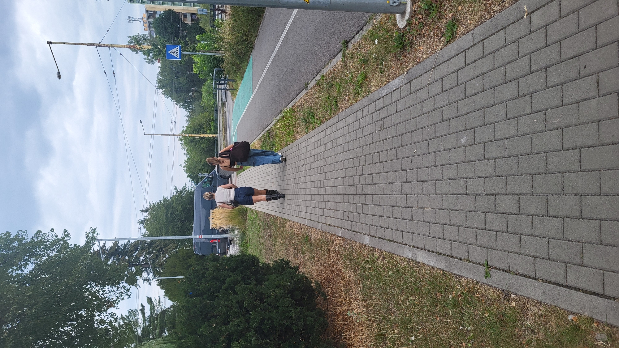 The image shows two women walking on a paved sidewalk next to a bike lane. The sidewalk is made of gray interlocking pavers. To the left of the sidewalk is a patch of dry grass and a dense green bush. In the background, a dark colored van is parked. A tall, rusted lamppost stands on the right side of the frame, near the bike lane. The bike lane has a green colored section. In the distance, there are trees and buildings, and the sky is overcast. The scene appears to be in an urban or suburban environment. Based on the presence of the "Predajňa elektro Bille" signage visible on a building in the background, the location is likely near the Billa supermarket on Vihorlatská Street in Prešov, Slovakia.